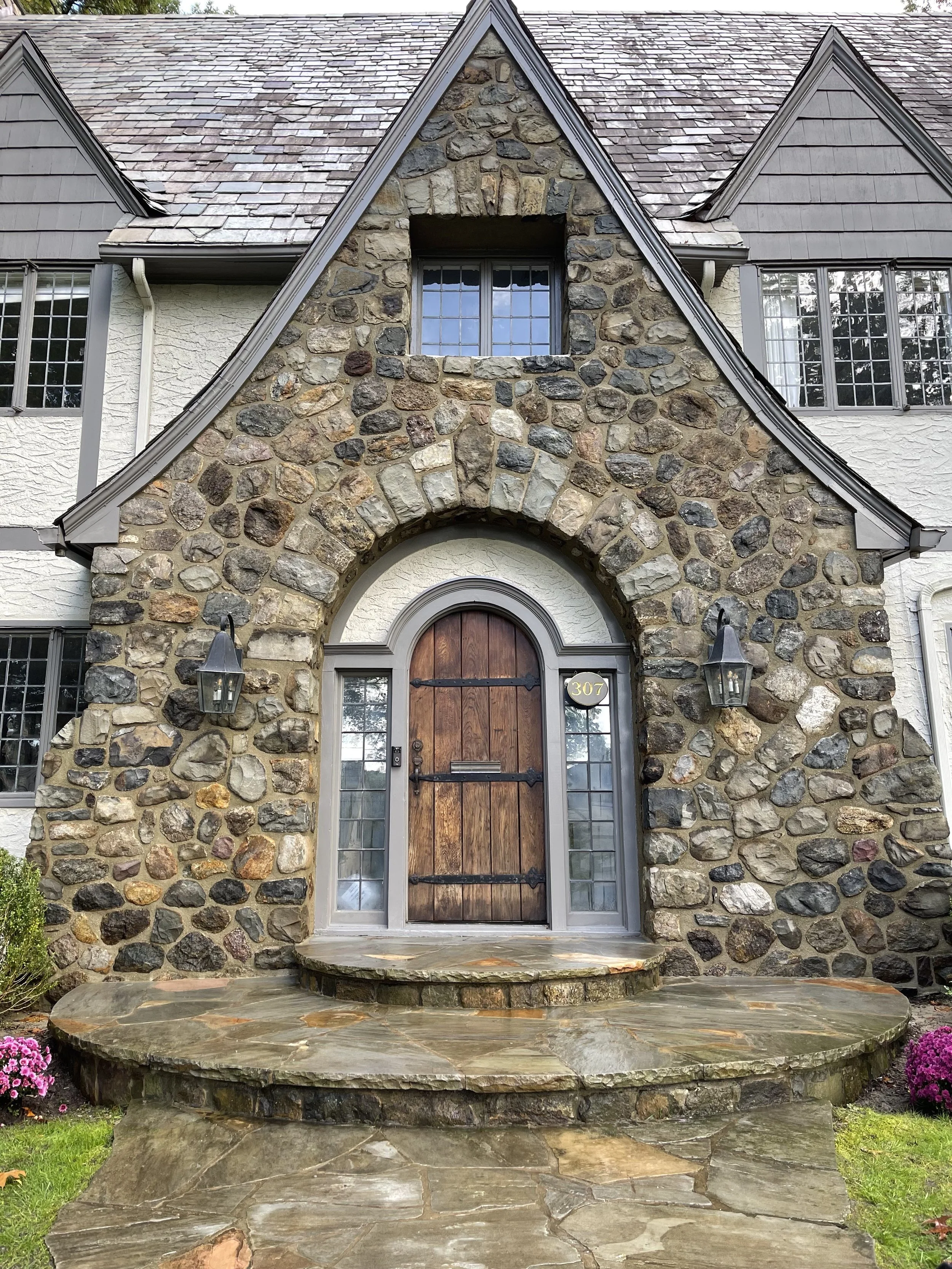 Front entrance of a house with a wooden door, stone archway, and stone steps, flanked by two black outdoor lanterns, with large windows on both sides of the door.
