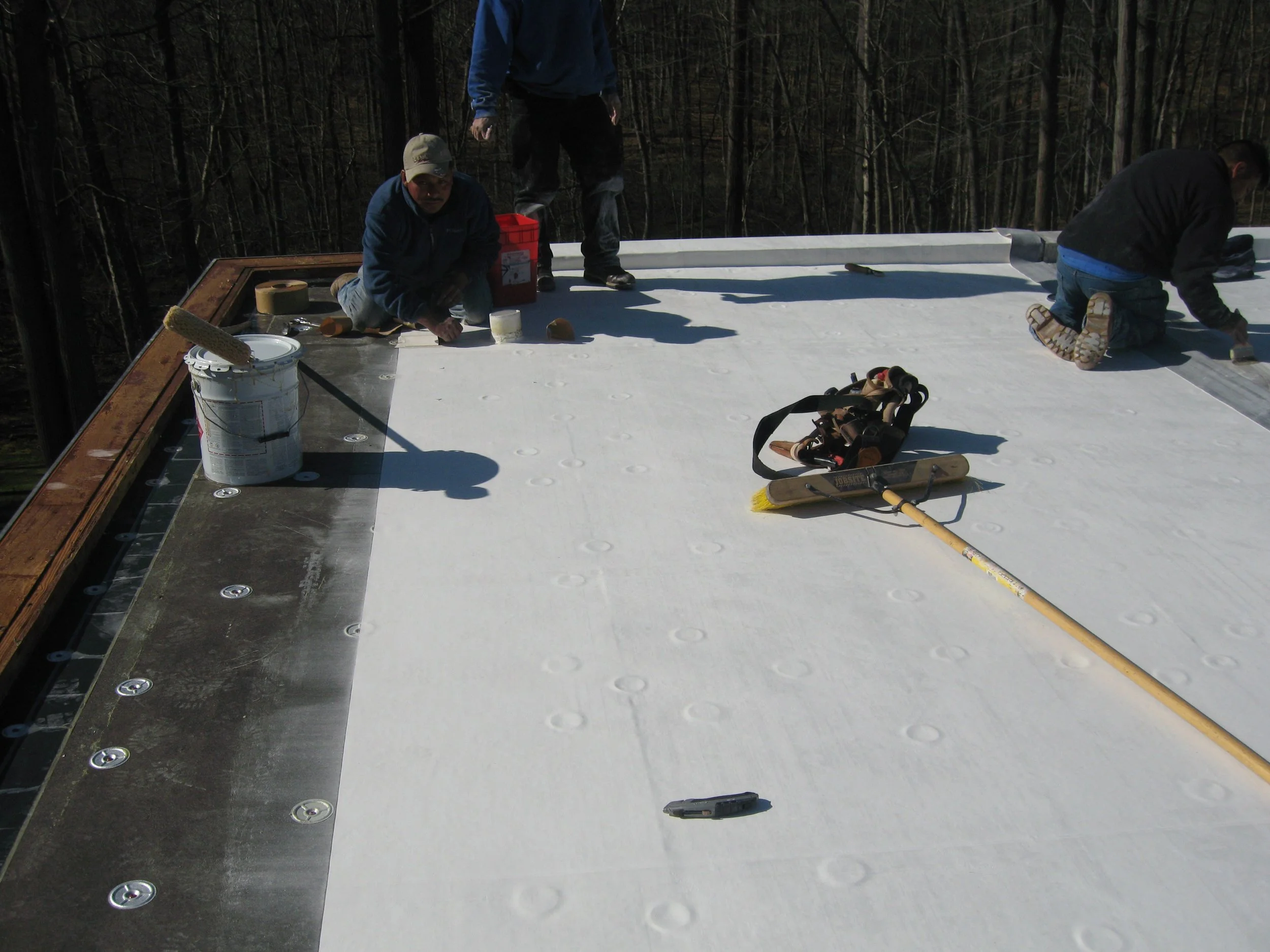 Workers installing or inspecting a white roofing material on a flat roof with tools and equipment nearby, surrounded by a wooded area.