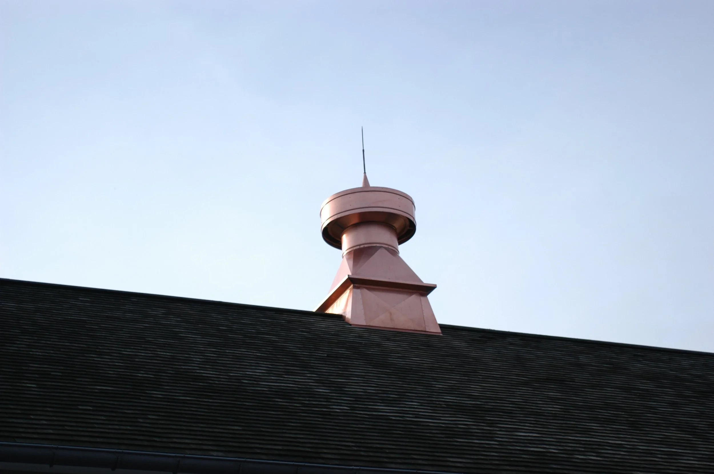 A copper-colored rooftop chimney with a spherical top and a thin antenna extending from it, set against a clear blue sky.