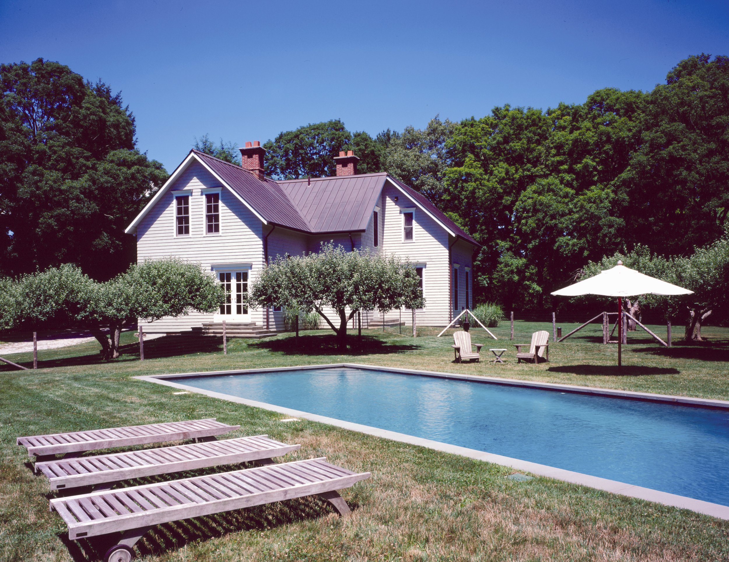 A backyard scene with a rectangular swimming pool, surrounded by green grass, with multiple wooden lounge chairs on the lawn. There are trees and a white house with a purple roof and chimneys in the background, under a clear blue sky.