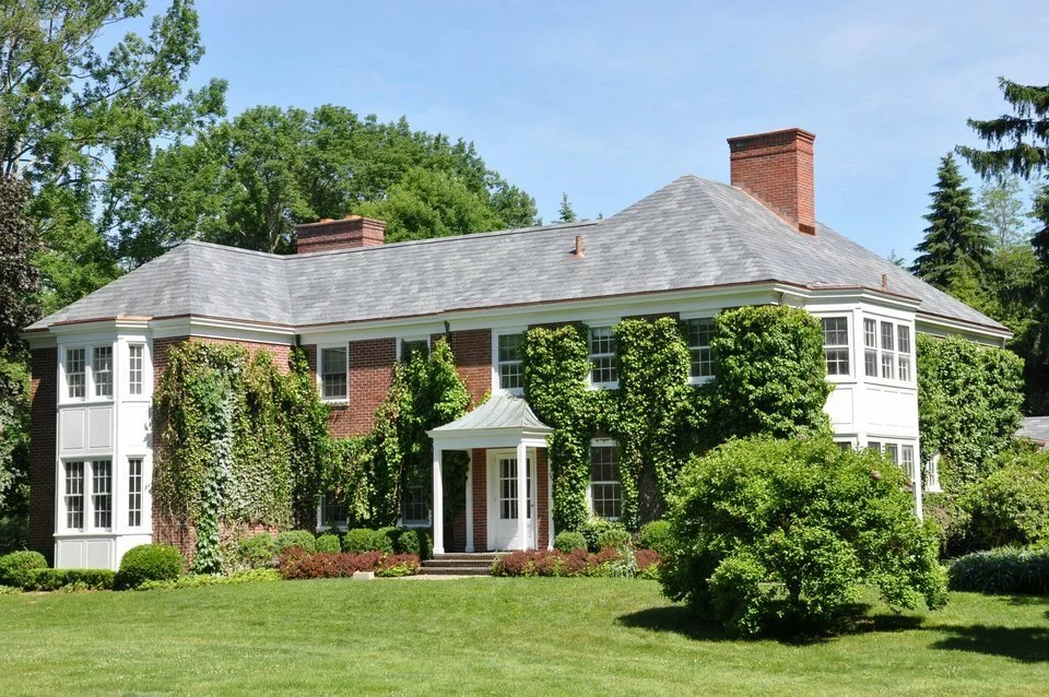 Large, historic brick house with white trim, surrounded by lush green trees and bushes, with a well-maintained lawn.