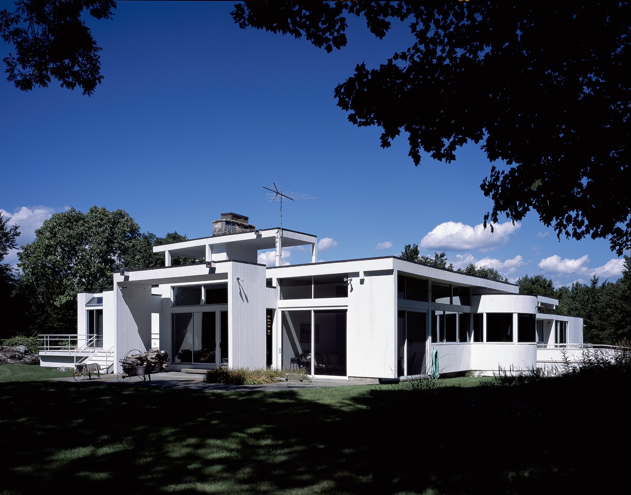 Modern white residential house with large windows and a flat roof, surrounded by a well-maintained lawn and trees, under a bright blue sky with some clouds.