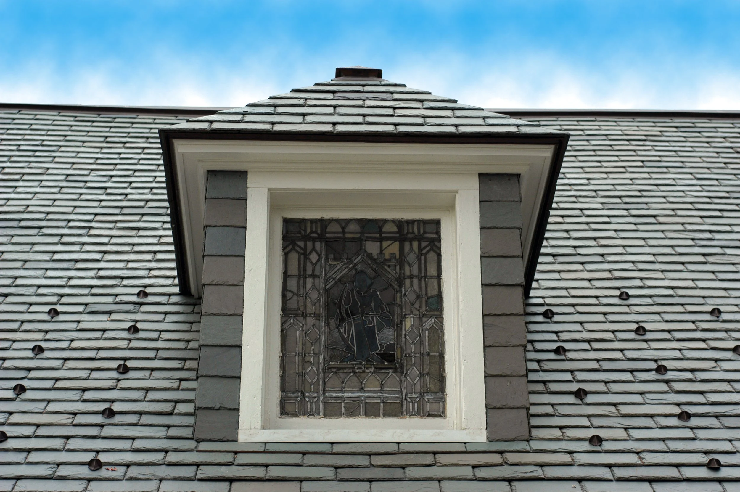 A close-up view of a dormer window on a house with a steep gray slate roof, featuring a stained glass window depicting a figure playing an instrument, set against a blue sky background.