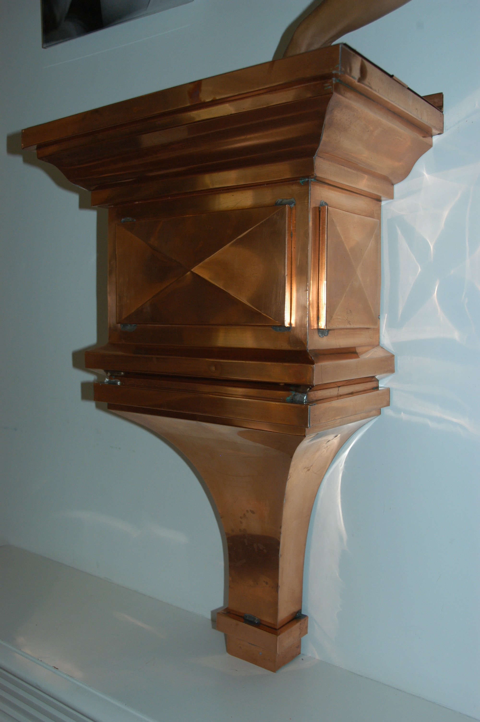 A wooden bar or countertop with a decorative uh landing or edge, supported by a curved wooden support structure, with a white wall and a white shelf or ledge in the background.