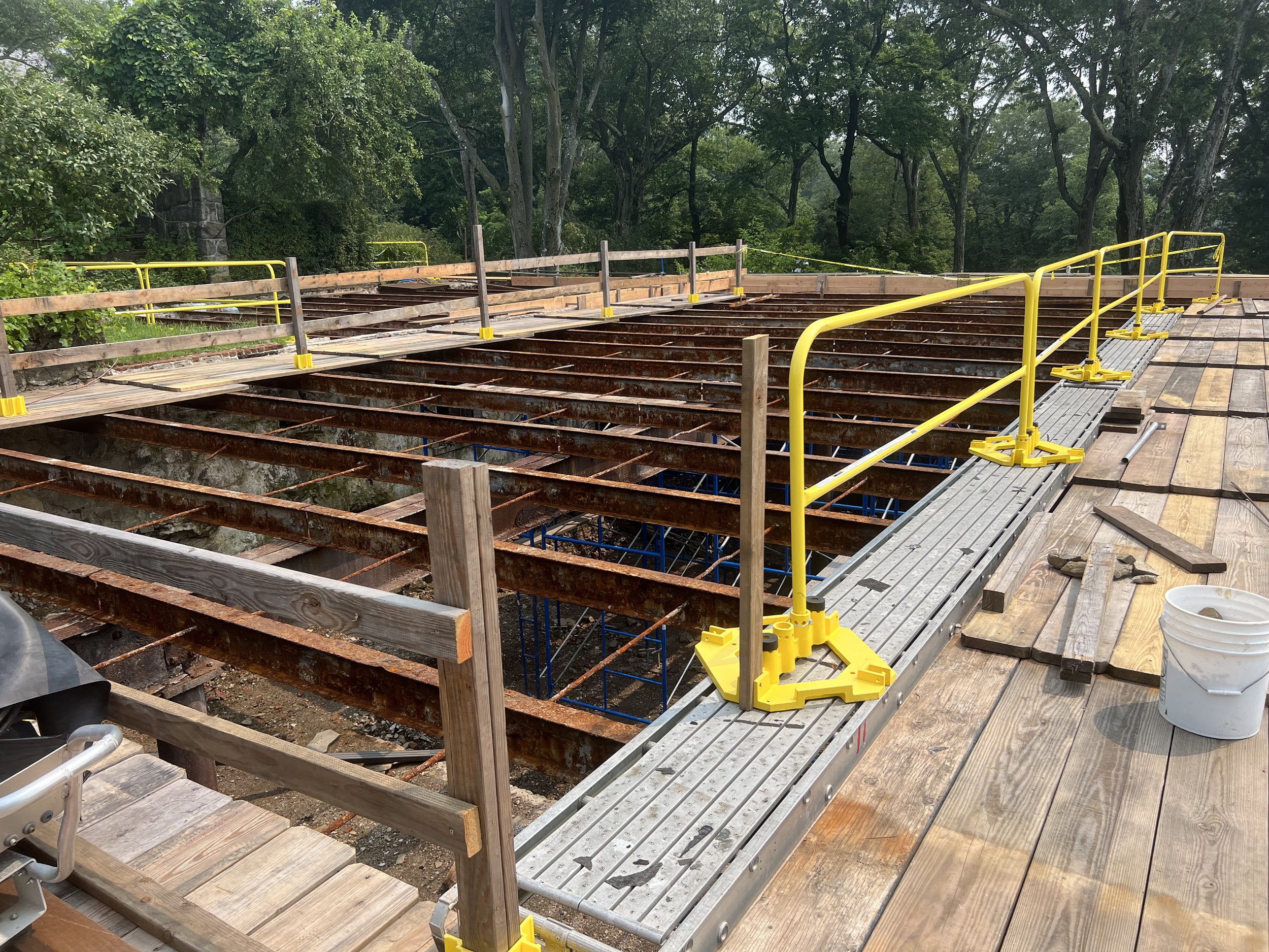 Construction site showing a partially built wooden and metal deck or platform with safety yellow railings, surrounded by trees and greenery.