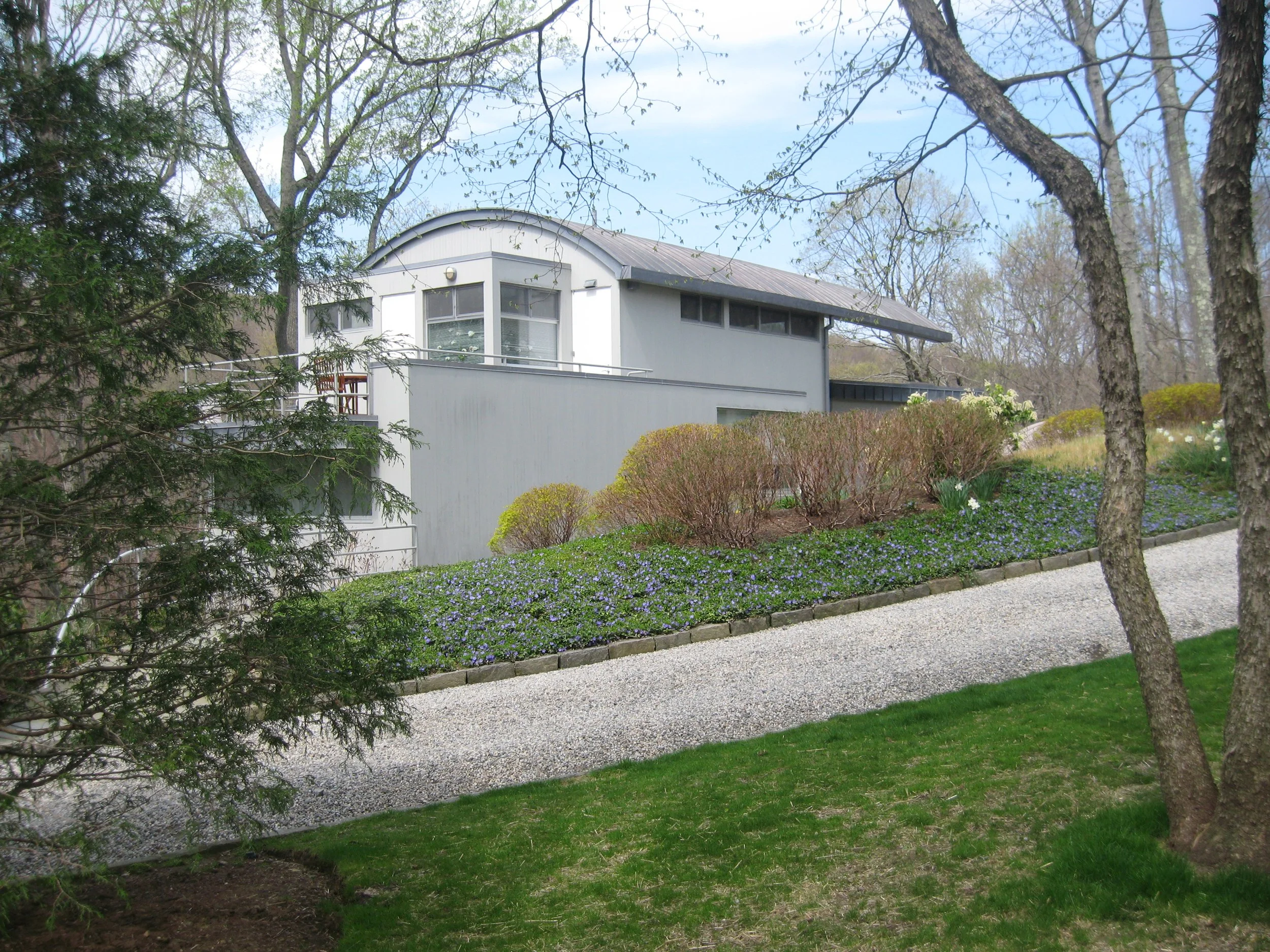 A modern white house with a curved roof seen through trees and bushes in a garden with a gravel path.
