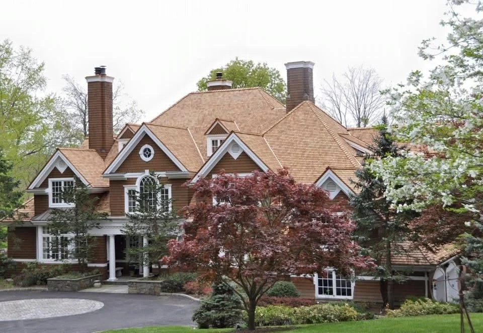A large, multi-story house with a brown wooden exterior and a complex roof with multiple gables, surrounded by trees and greenery.