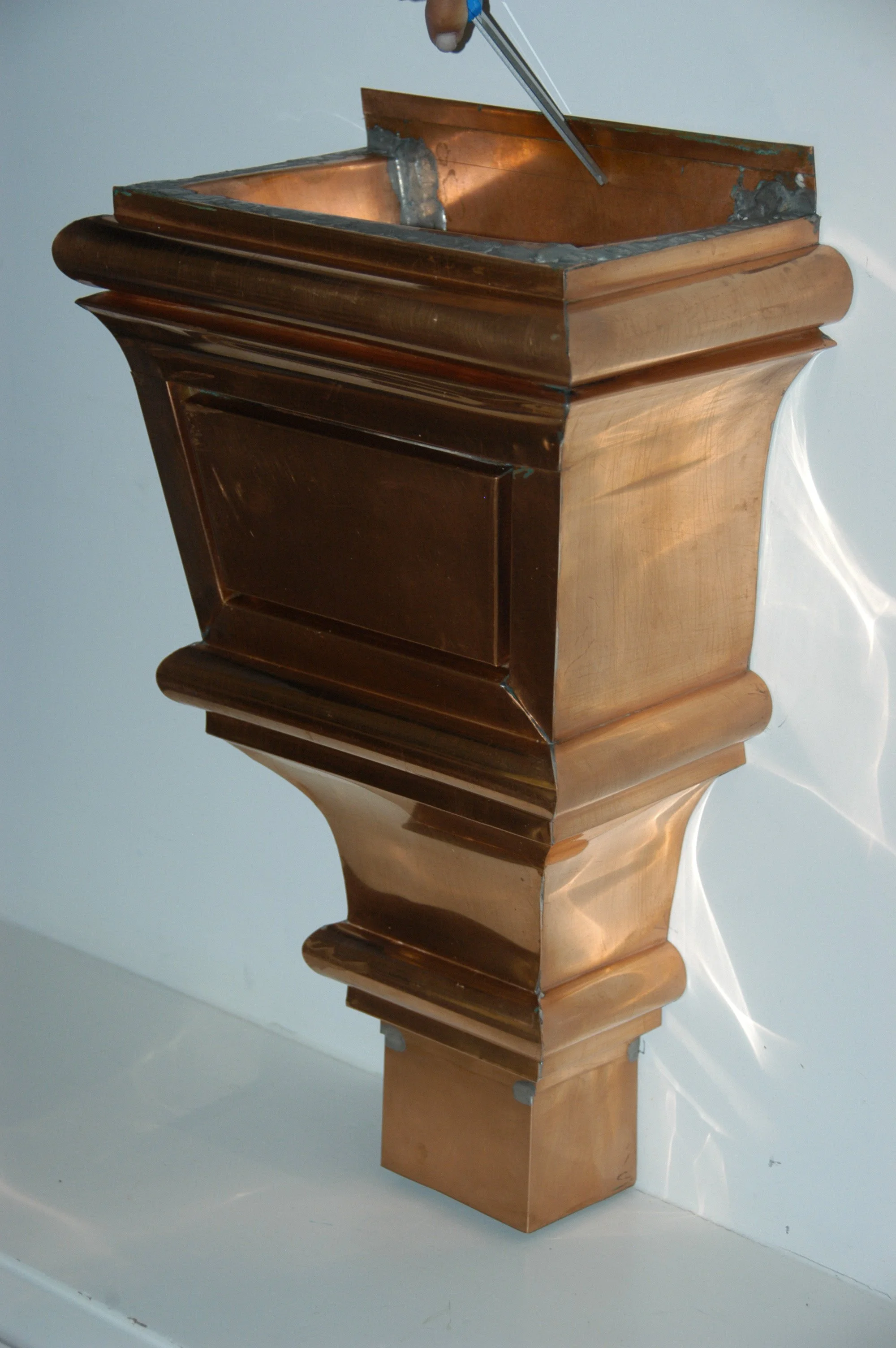 Wooden furniture piece with curved decorative edges, placed against a light-colored wall, with a visible screw or bolt at the top corner. The perspective is from below, showing the detailed craftsmanship of the furniture's design.