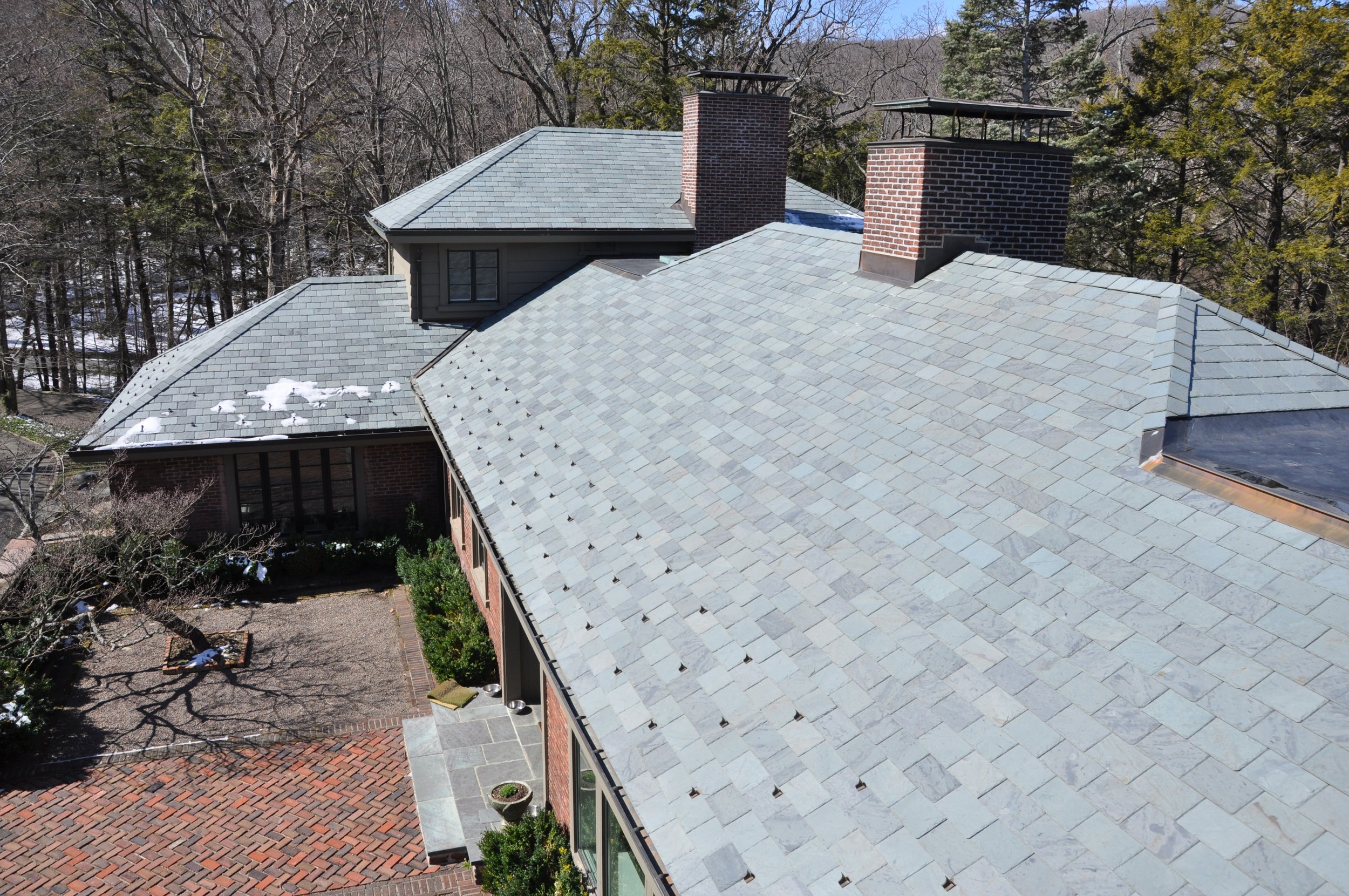 Aerial view of a house's rooftop with light gray shingles, two brick chimneys, and snow patches, surrounded by leafless trees and a wooded background.