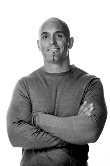 Black and white portrait of a bald man with a goatee, wearing a long-sleeve shirt, crossing his arms and smiling.