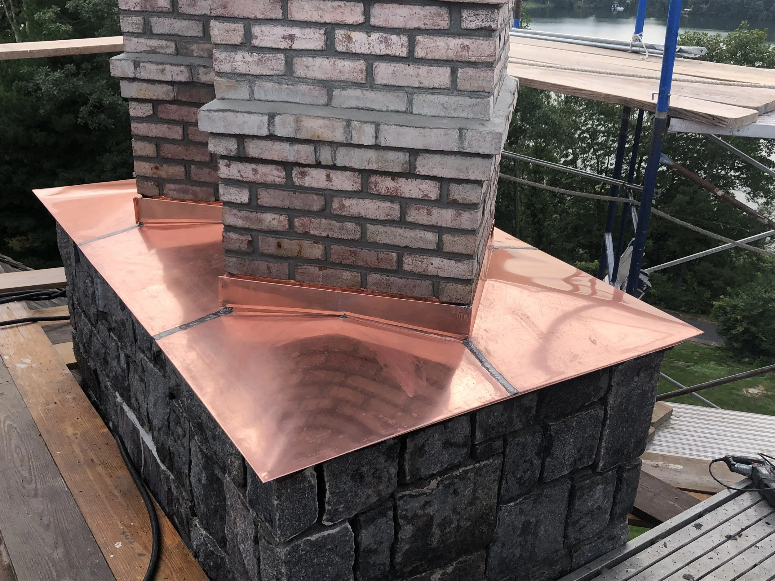 A brick chimney on a building under construction with copper flashing at the base, a wooden platform, and blue scaffolding in the background.