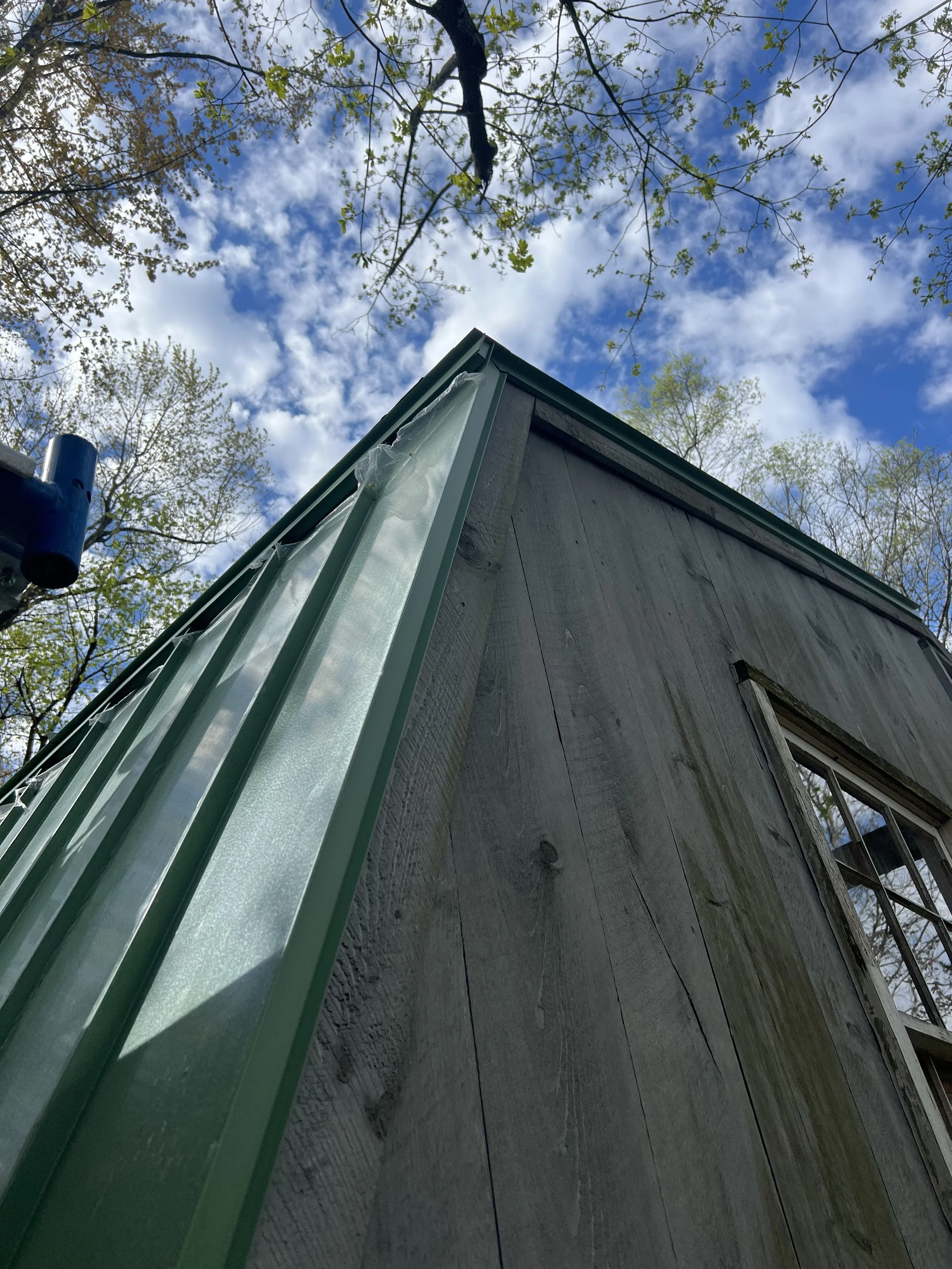 Low-angle view of a house corner with green trim, a section of the wall, and a gutter, set against a backdrop of blue sky with scattered clouds and tree branches with budding green leaves.