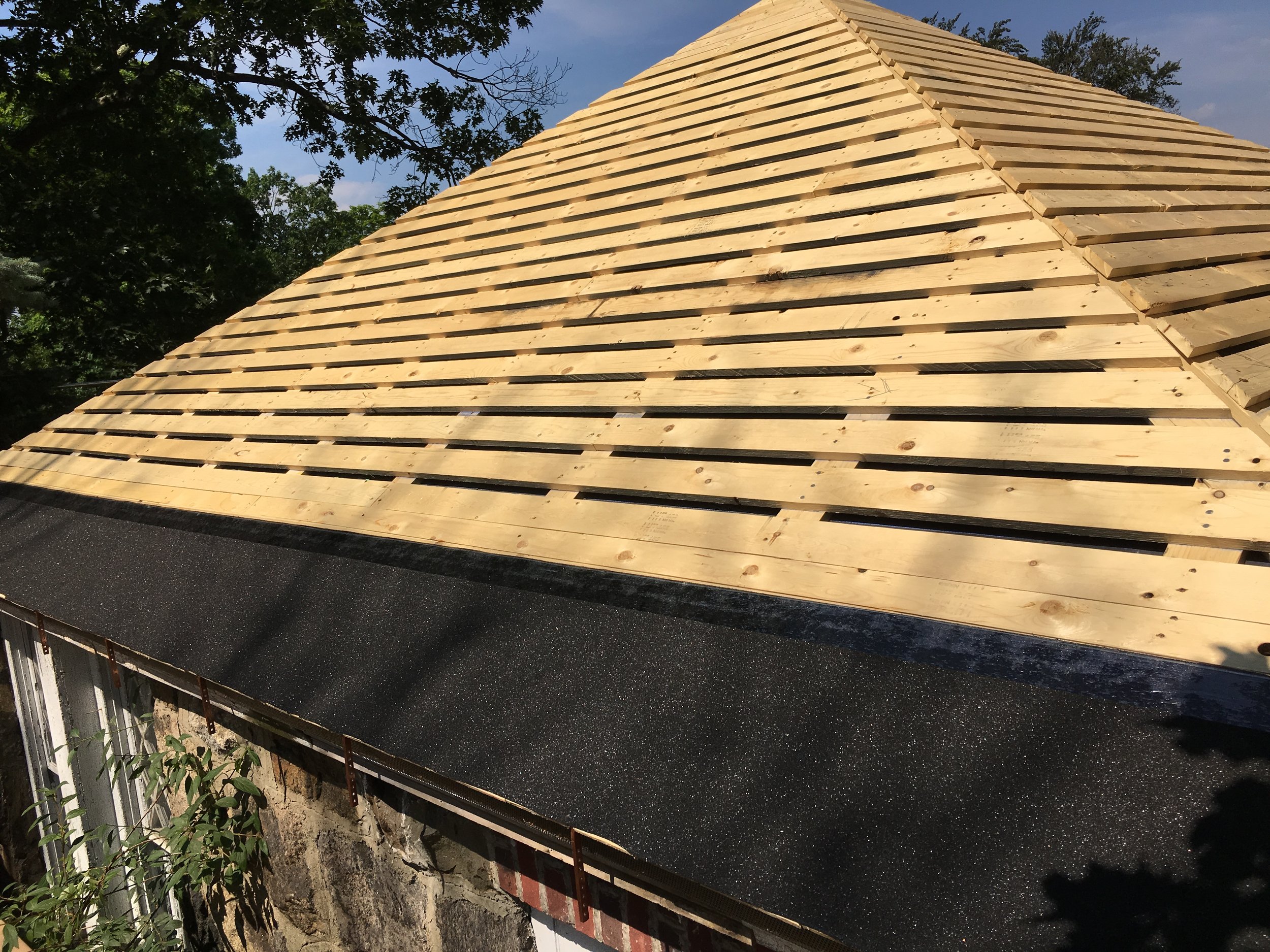 Side view of a building with a roof under construction, showing wooden roof trusses and dark roofing material partially installed.