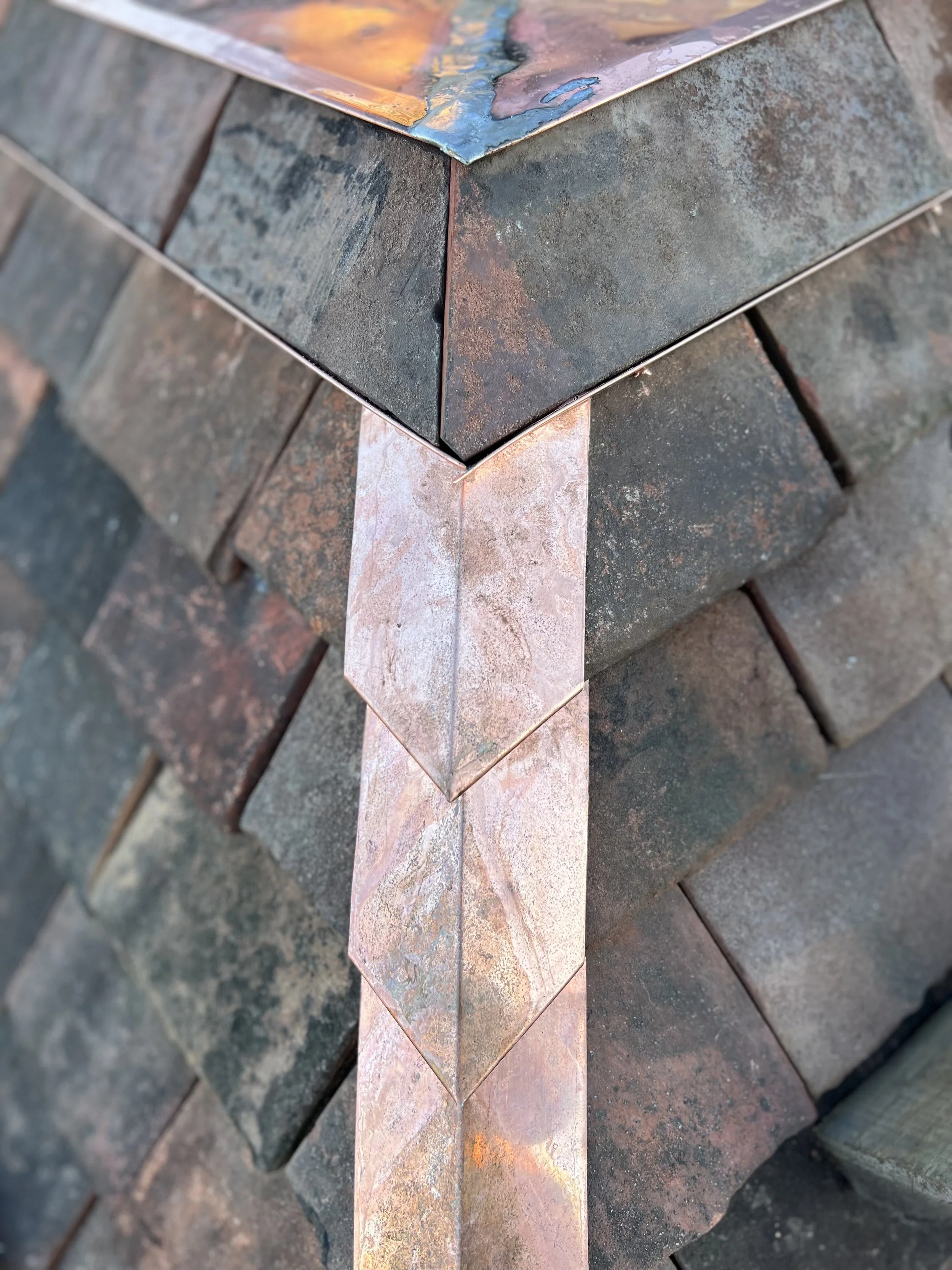 Close-up of a corner of a metal sculpture with colorful, textured tiles arranged in a diamond pattern, placed on a brick surface.