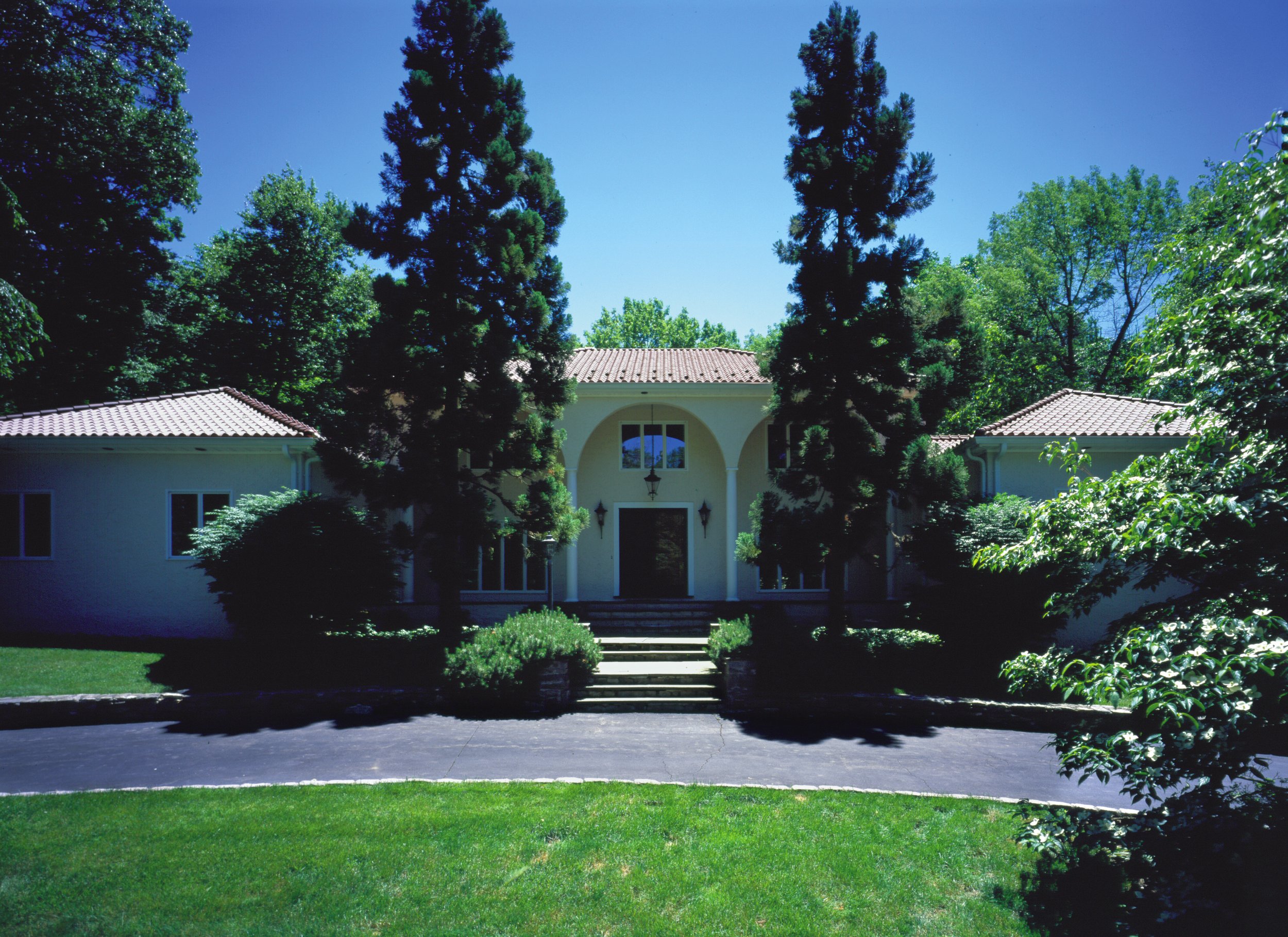 Large house with white walls and a red tile roof, surrounded by tall trees and lush greenery, blue sky overhead.
