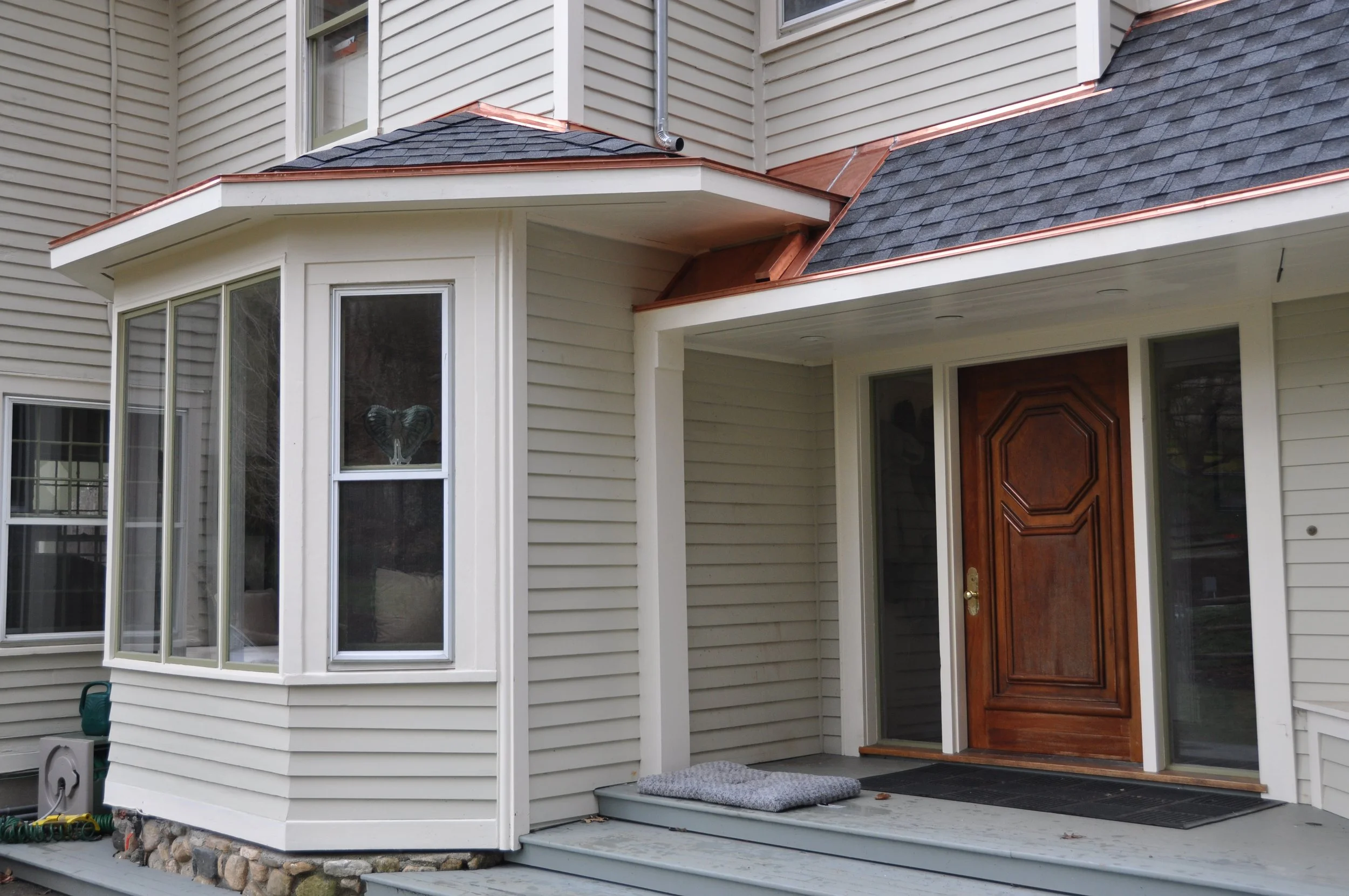 Close-up of the front porch and entrance of a house with beige siding, a wooden front door, and a small steps leading up to the door, with a gray doormat and a gray rug.