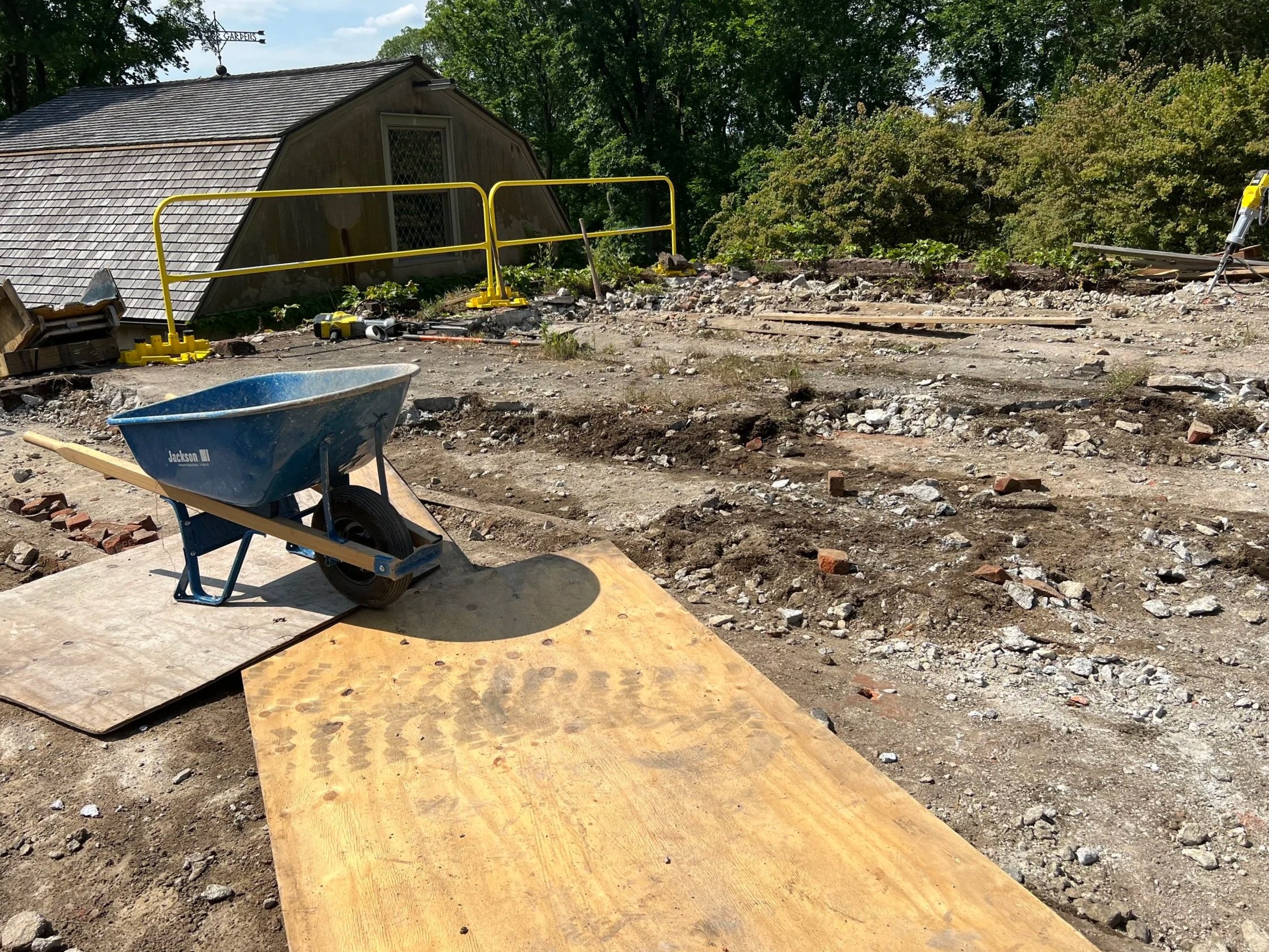 A construction site with a wheelbarrow, plywood, and various tools scattered across uneven dirt ground. A small wooden building with a shingled roof is visible in the background, surrounded by green trees.