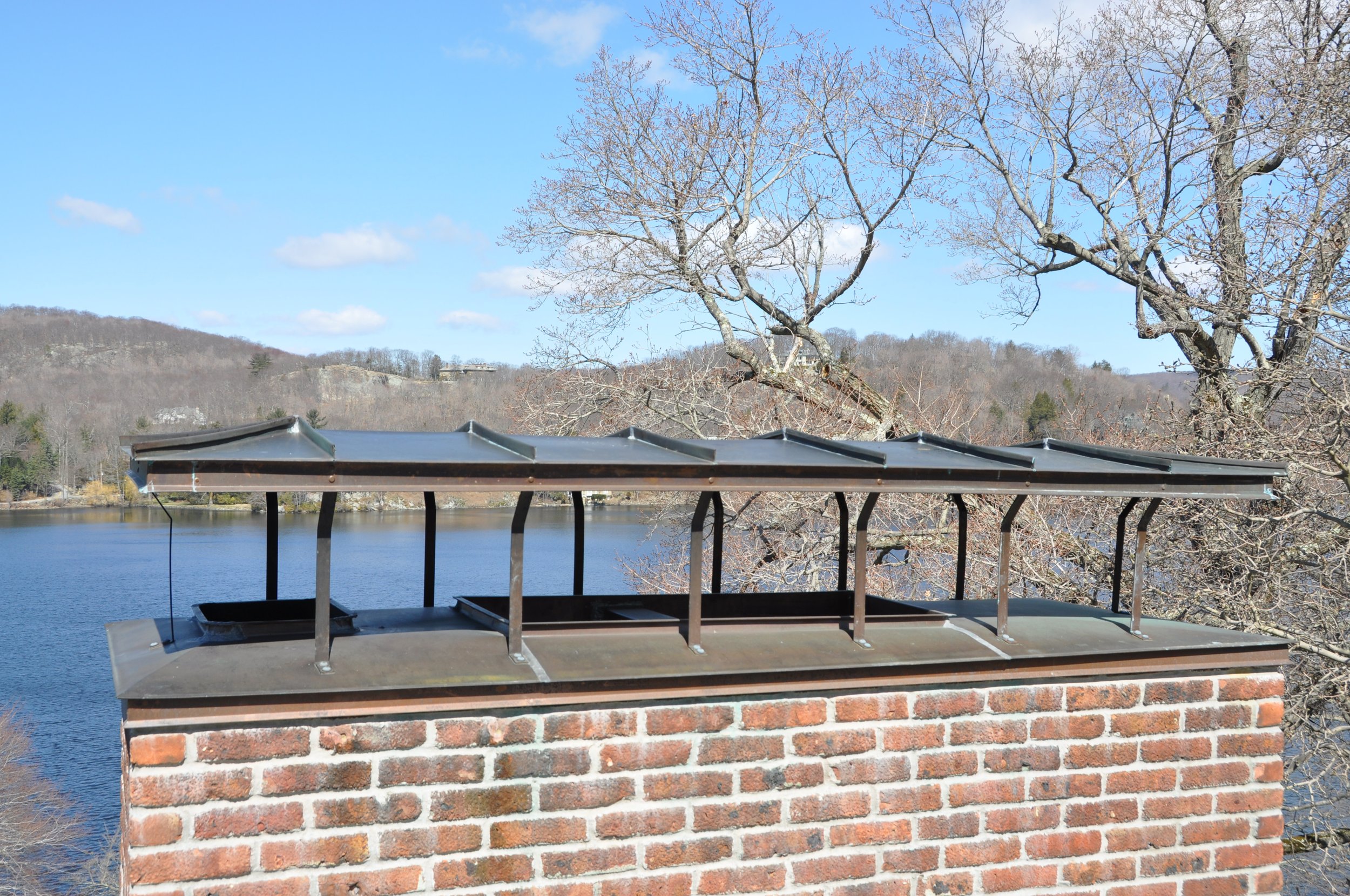 A brick chimney with a metal roof cap and a rope hanging from one corner, set against a backdrop of a lake, leafless tree branches, and distant hills under a partly cloudy blue sky.
