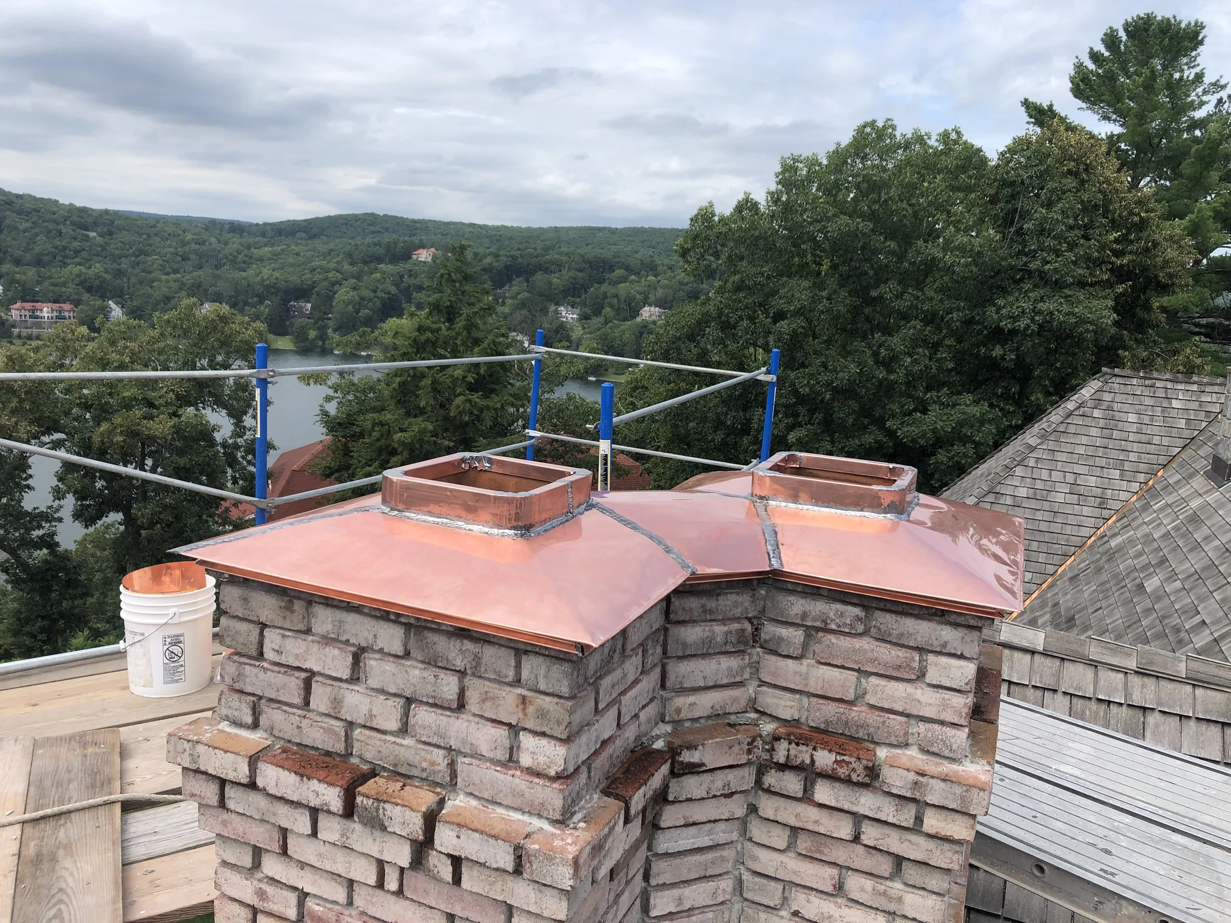 A construction site featuring a brick chimney with a copper cap, scaffolding around it, and a bucket nearby. The background shows a scenic view of trees, houses, and a lake under a cloudy sky.