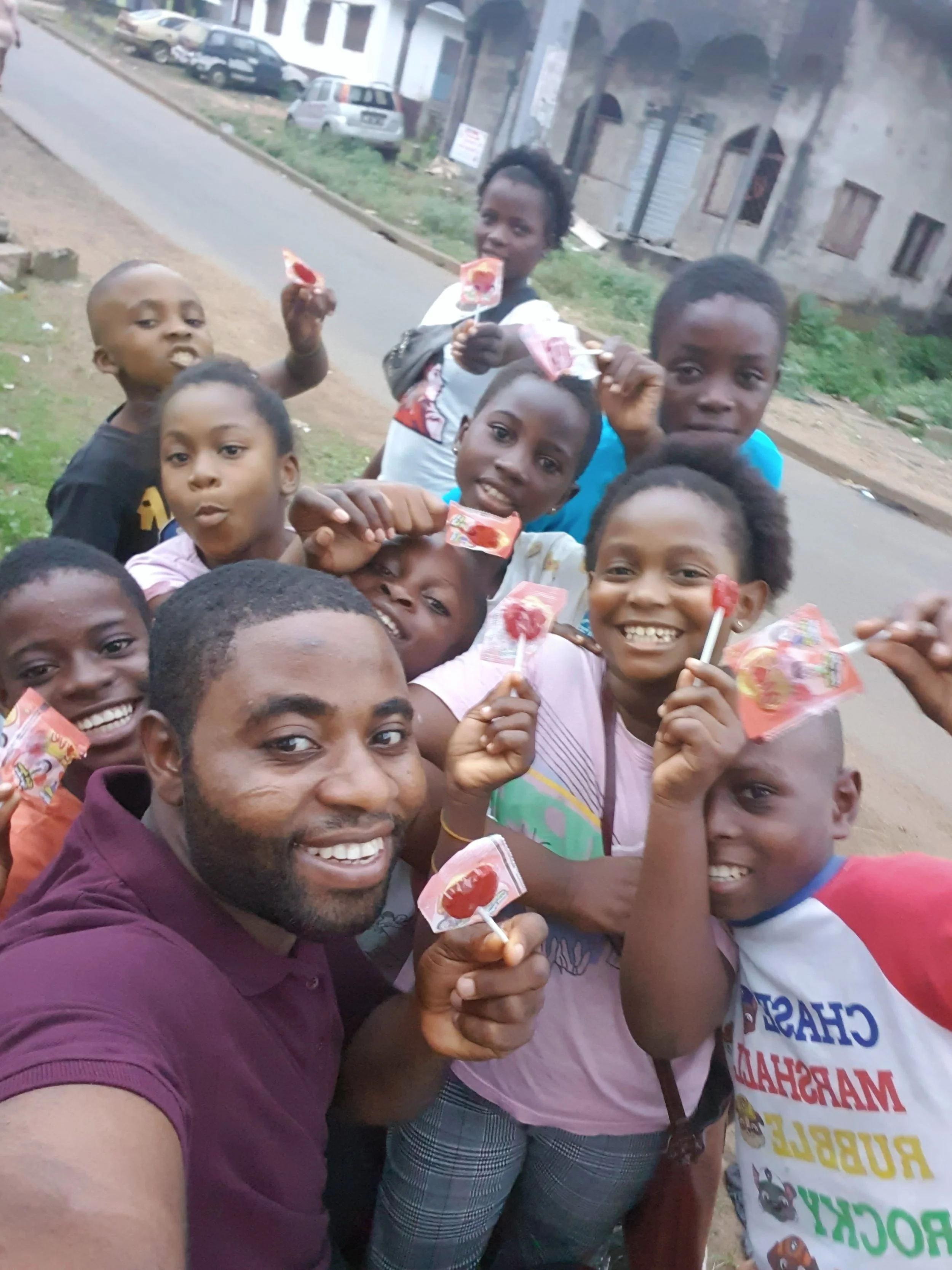 A group of children and a man are smiling and holding up lollipops outside on a street. There are houses and parked cars in the background.