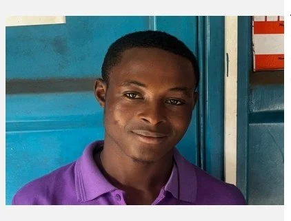 A young man with dark skin and short black hair, wearing a purple polo shirt, standing indoors in front of a blue wall and door.