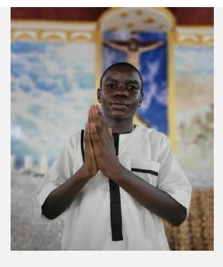 Young man in a white shirt standing for prayer or blessing inside a church with a decorative background and an image of Jesus in the background.
