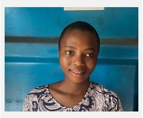 A young woman with short hair and a patterned top stands against a blue wall, smiling at the camera.