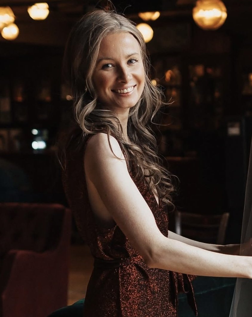A smiling woman with long, wavy hair wearing a sleeveless, sparkly dress in a dimly lit indoor setting with warm lighting.