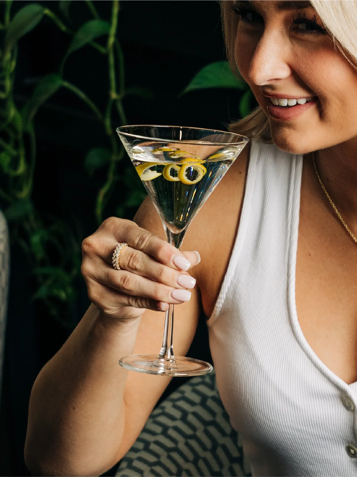 Close-up of a woman holding a martini glass with a lemon twist garnish, smiling, wearing a white sleeveless top, with green plants in the background.
