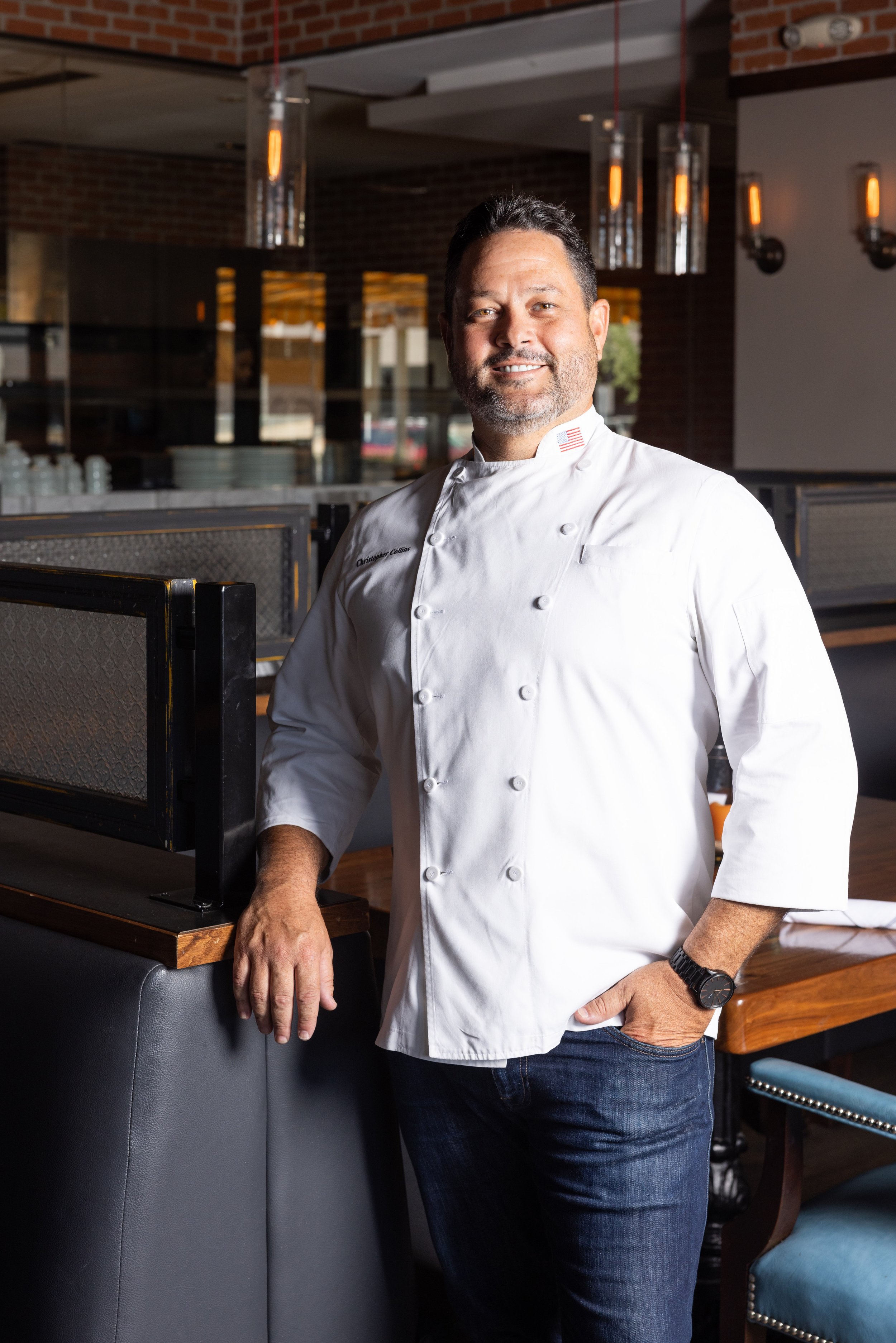 A male chef standing in a restaurant, wearing a white chef coat with an American flag on the collar. He has short dark hair, a beard, and is smiling. The background shows a modern restaurant interior with brick walls, pendant lighting, and tables with chairs.
