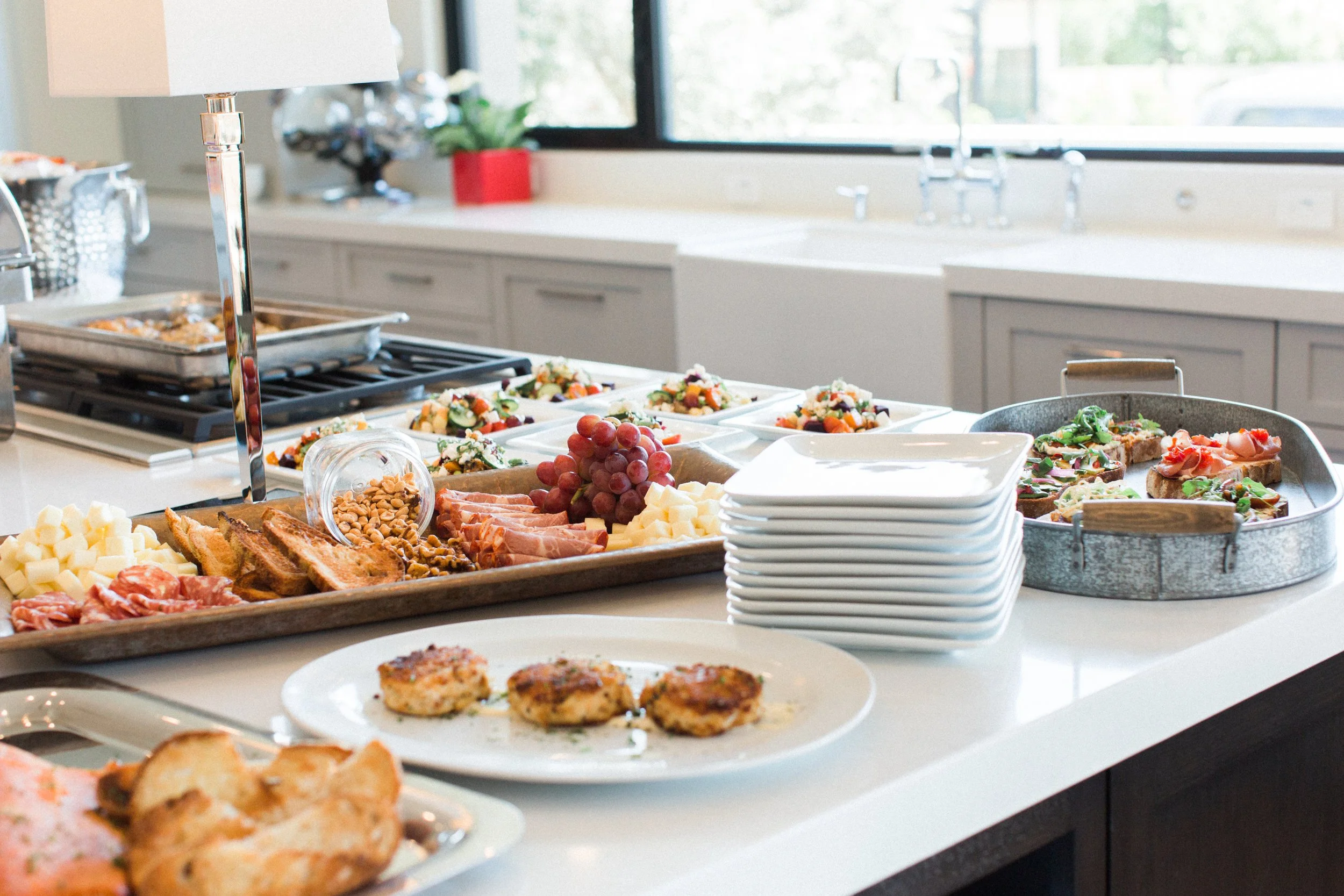 A spread of appetizers and finger foods on a kitchen countertop, including cheese, crackers, grapes, sliced meats, salads, and small sandwiches, with a window and kitchen sink in the background.