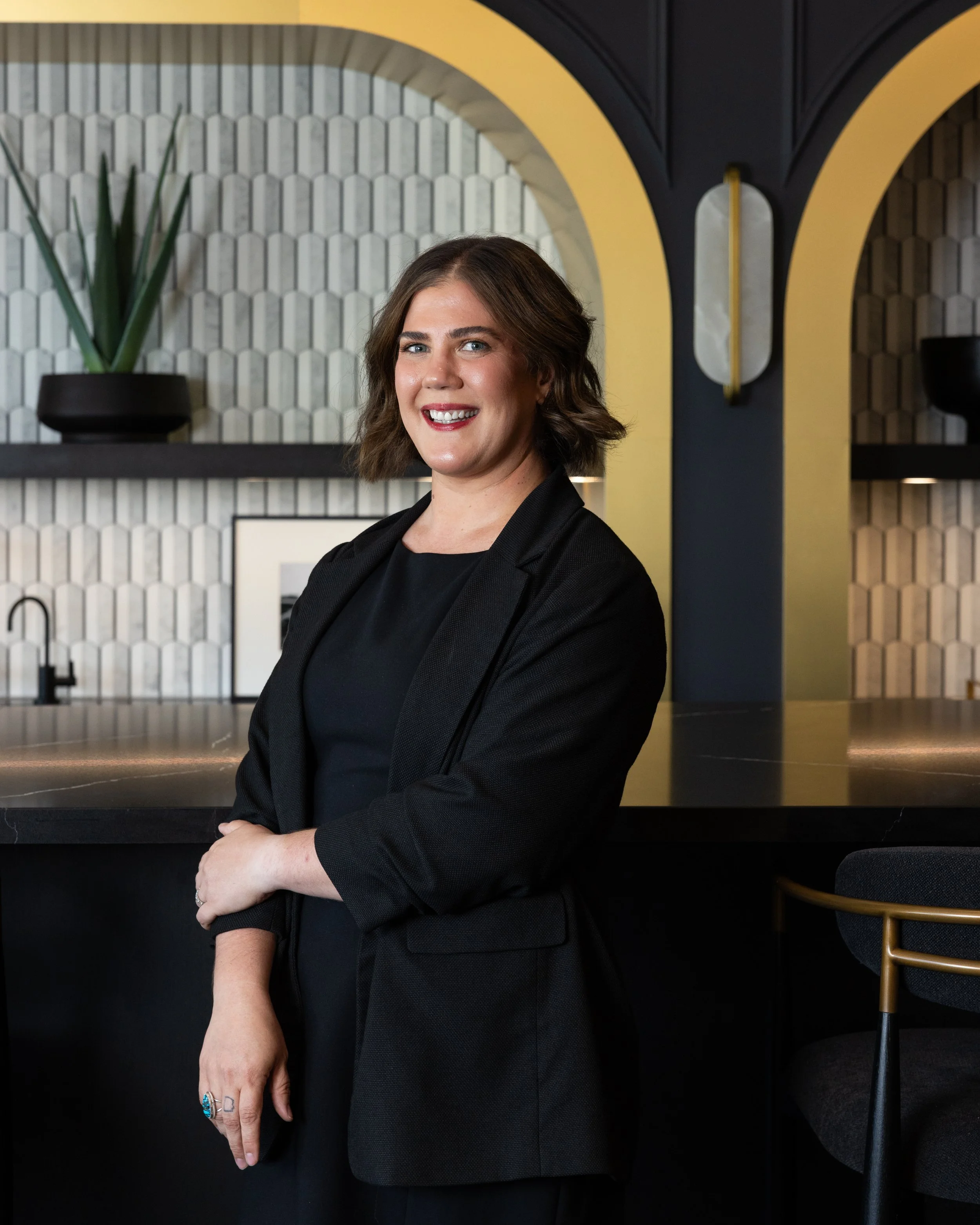 Portrait of a woman with short brown hair, wearing a black blazer, standing in a modern interior with textured wall tiles, minimalist decor, and a decorative plant.