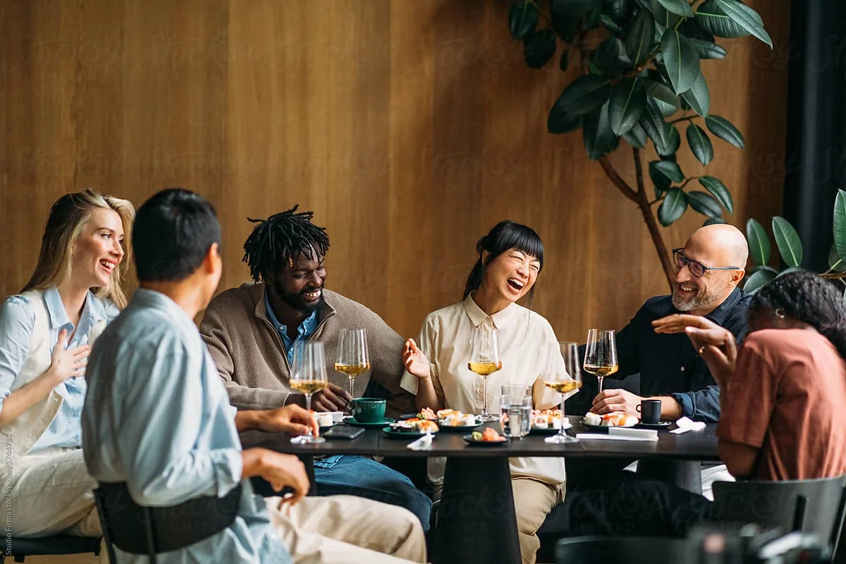 Group of six diverse people enjoying drinks and laughing together around a dining table in a well-lit room with wood-paneled walls and large green plants.