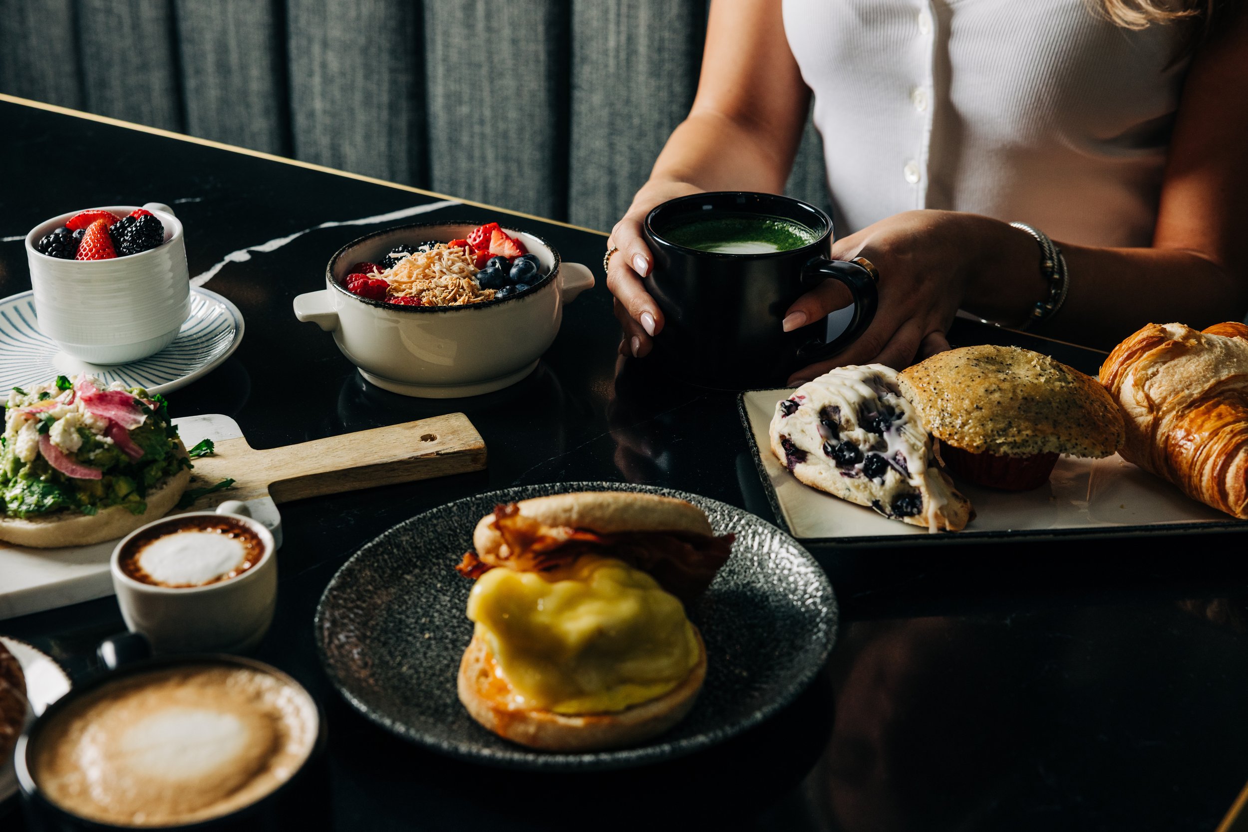A table with various breakfast foods including a bowl of mixed berries, a plate of scones with clotted cream and jam, a cup of coffee, a muffin with lemon curd, and a person holding a black mug of green smoothie.