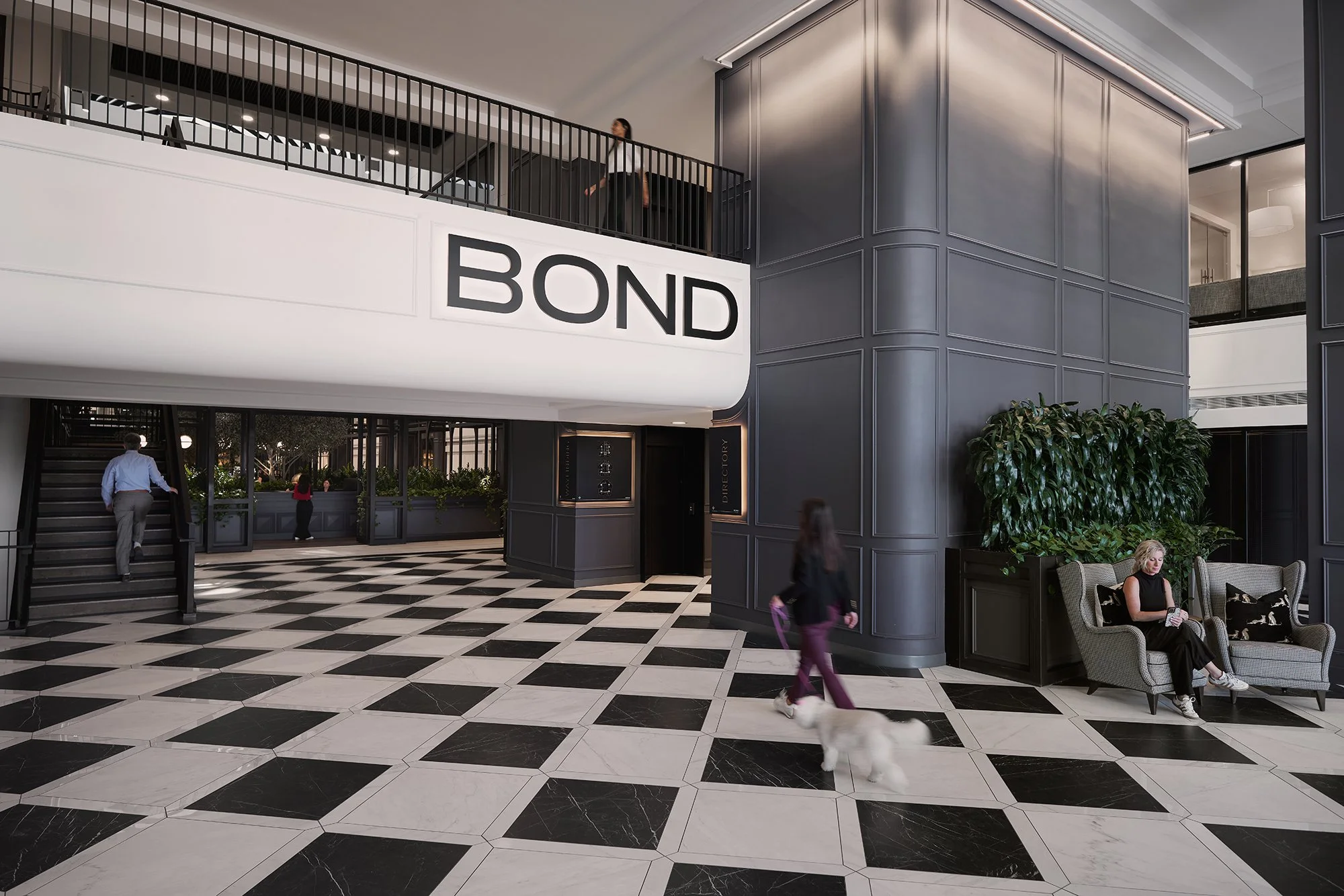 Interior of a modern building with black and white checkered floor, a large white sign with black letters spelling 'BOND', multiple people walking and sitting, and a woman with a dog sitting on a sofa.