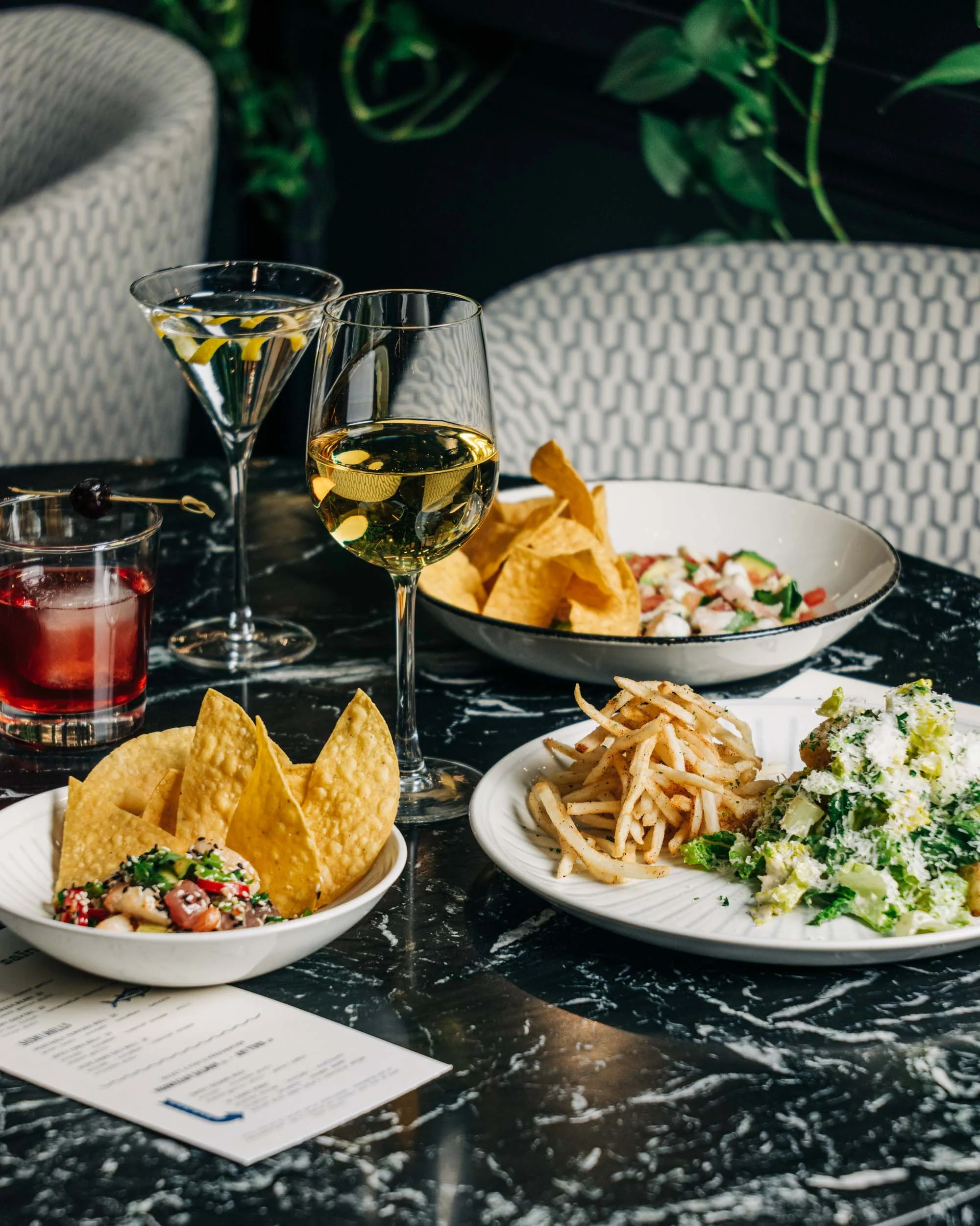Table set with various foods and drinks, including a glass of white wine, a martini with lemon peel, a glass of red drink, a bowl of chips, a salad with vegetables, and a plate of pasta. There are chairs and green plants in the background.