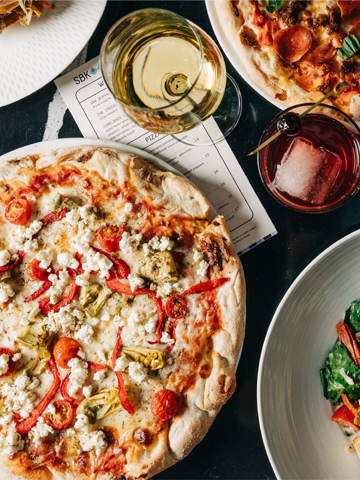 A table scene with a pizza topped with tomatoes, red peppers, artichokes, and cheese, a glass of white wine, a glass of red drink with a cherry, and a salad on a white bowl.