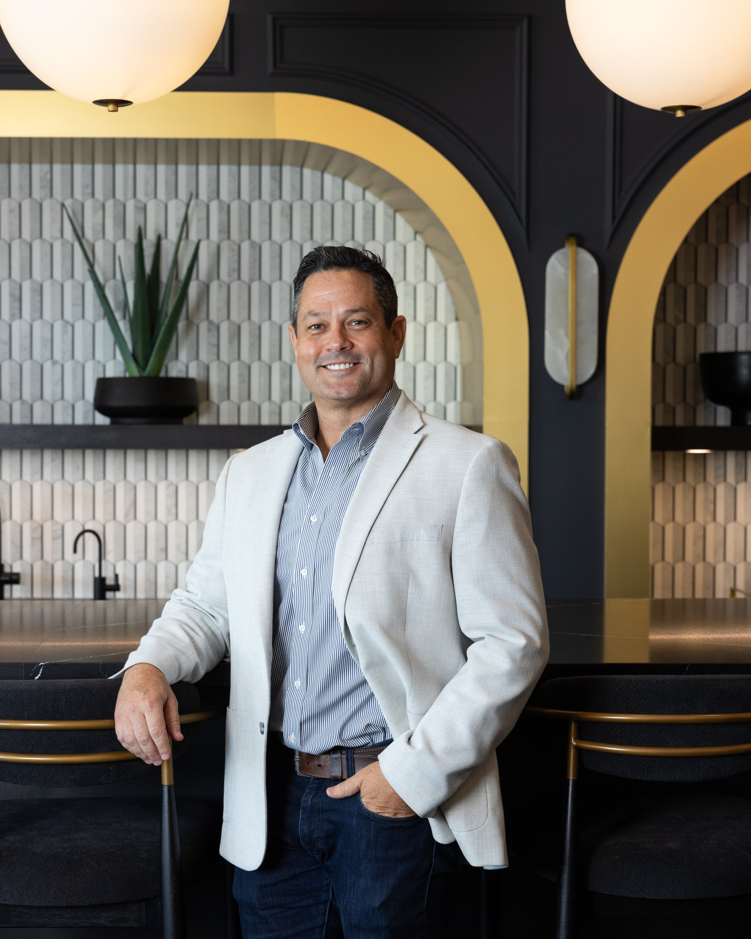A man in a light-colored blazer and jeans standing in front of a modern interior with black and yellow accents, decorative plants, and geometric wall patterns.