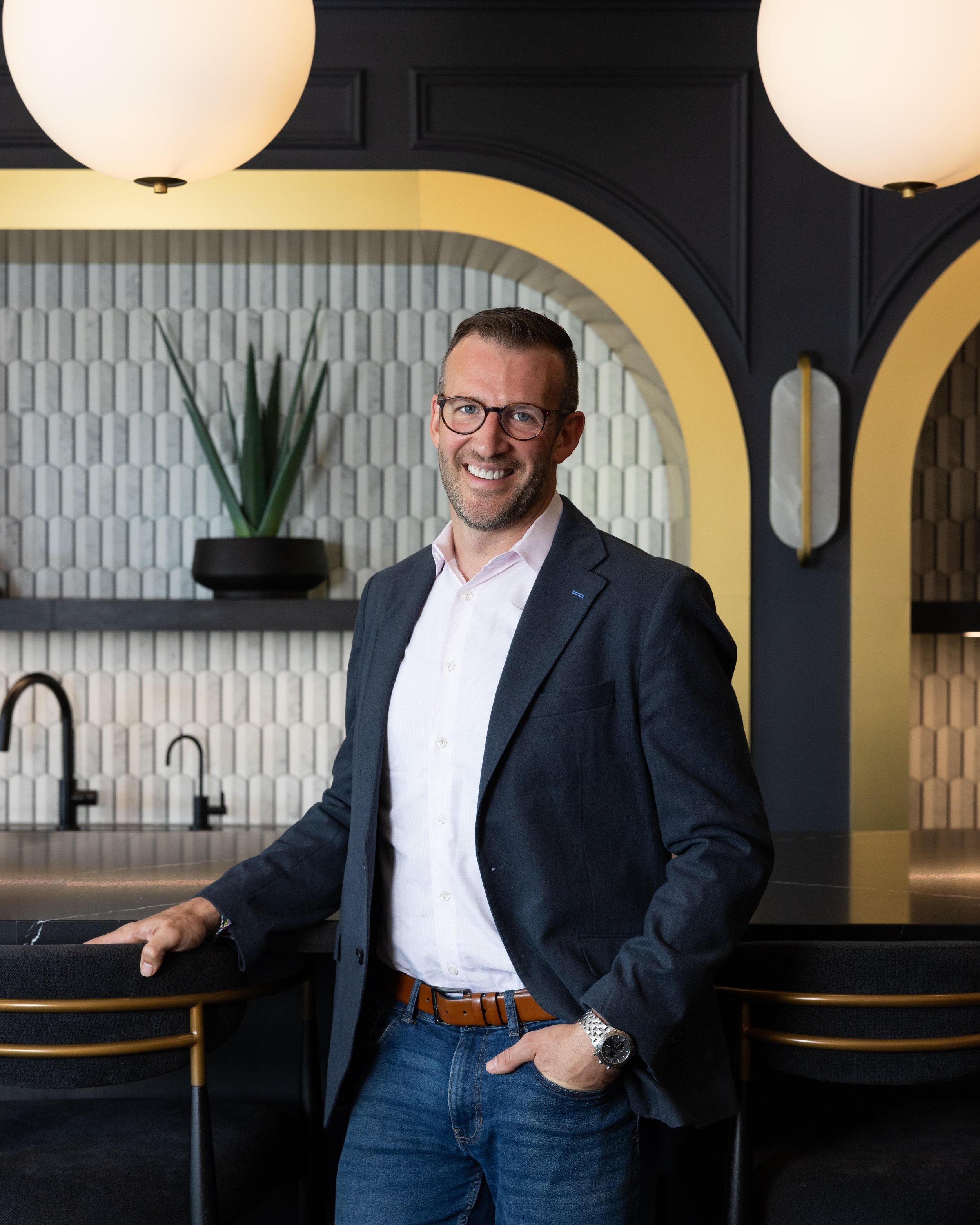 A smiling man in a blazer, white shirt, and jeans, wearing glasses, standing in a modern, stylish room with a decorative tiled wall, yellow arch, black pendant lights, a plant, and a black counter.