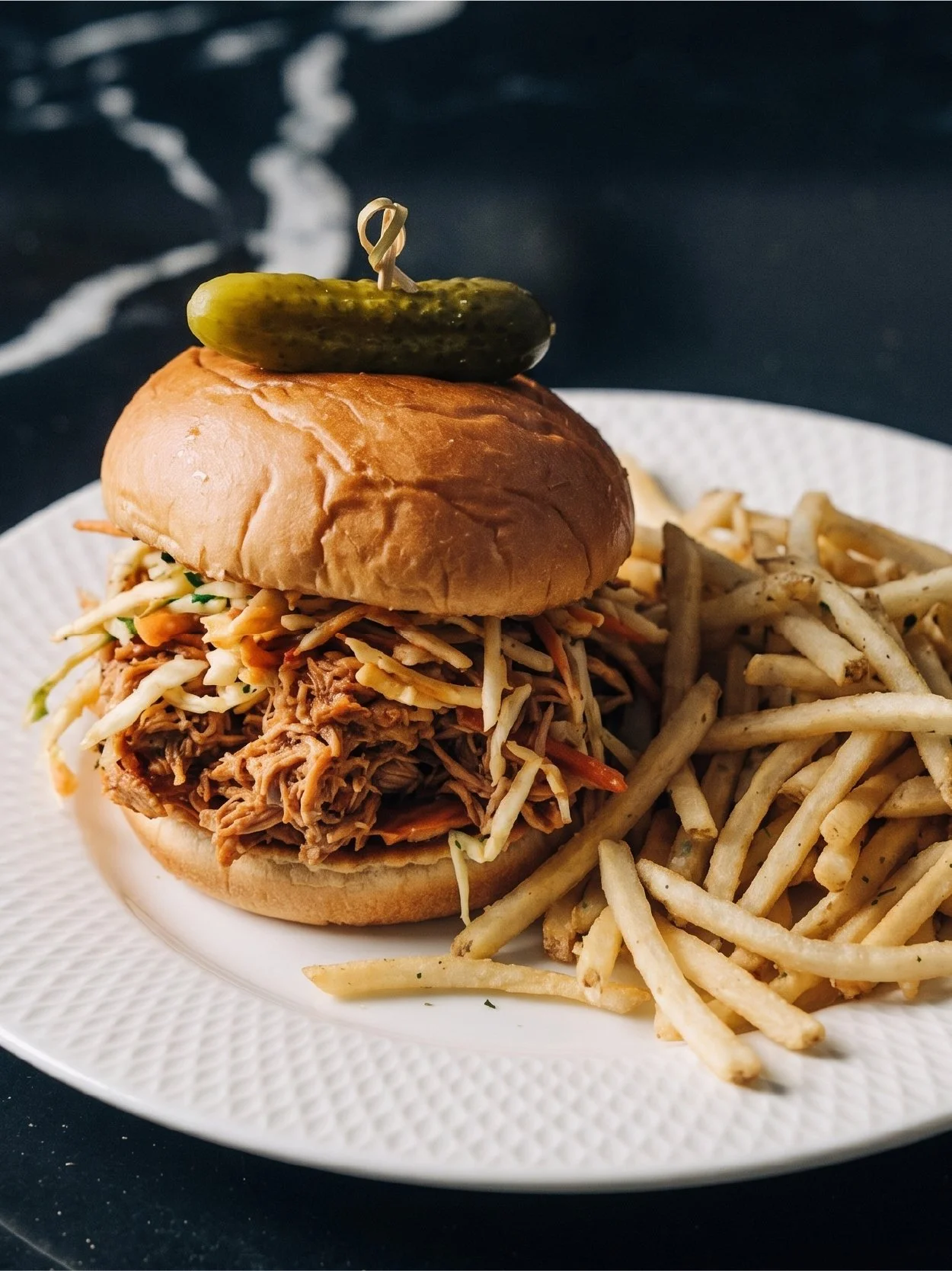 Pulled pork sandwich with coleslaw on a bun, topped with a pickle, served with French fries on a white plate.