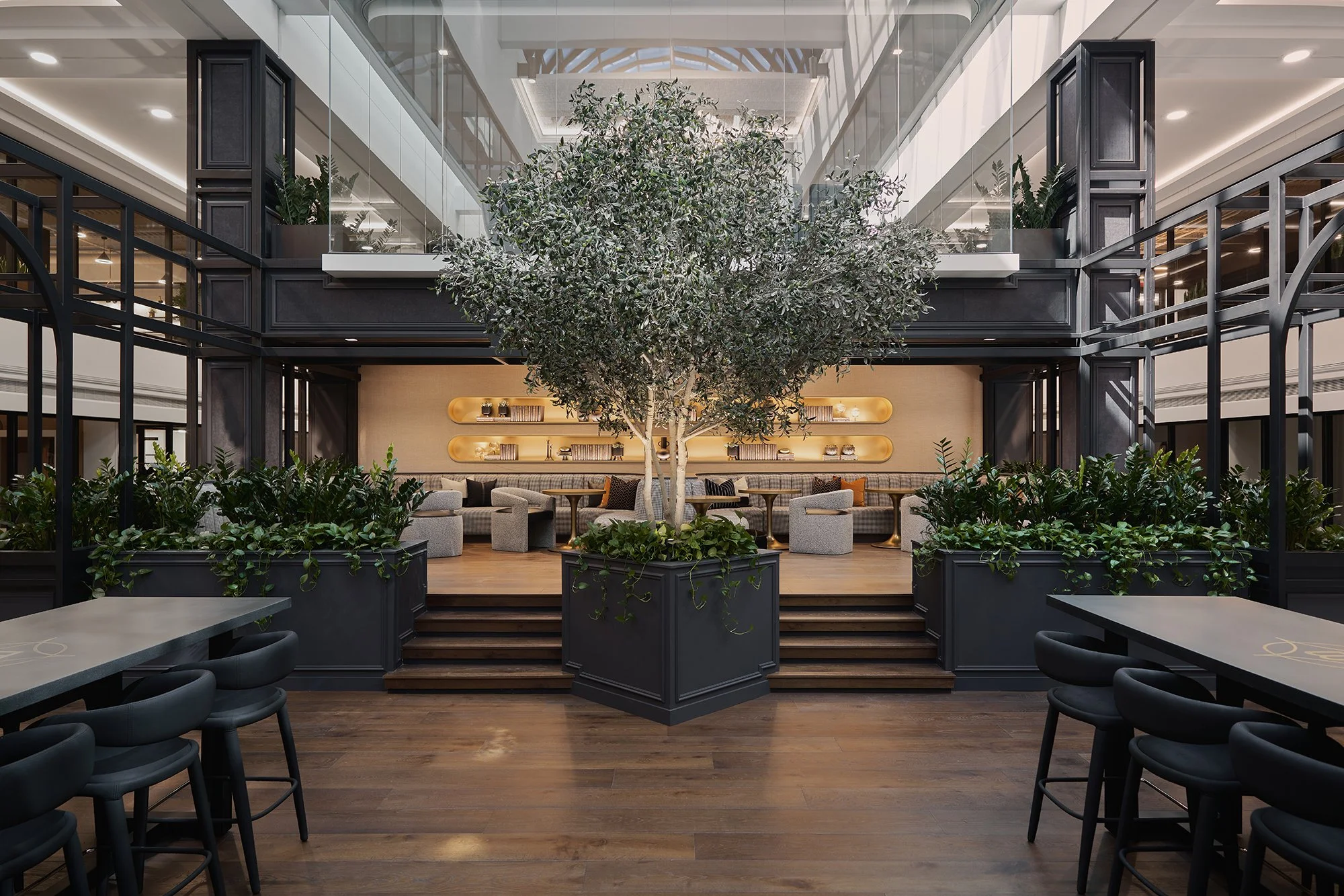 Interior of a modern atrium with a large central tree, surrounding seating area with armchairs and tables, black framing, and an upper level with glass walls and potted plants.