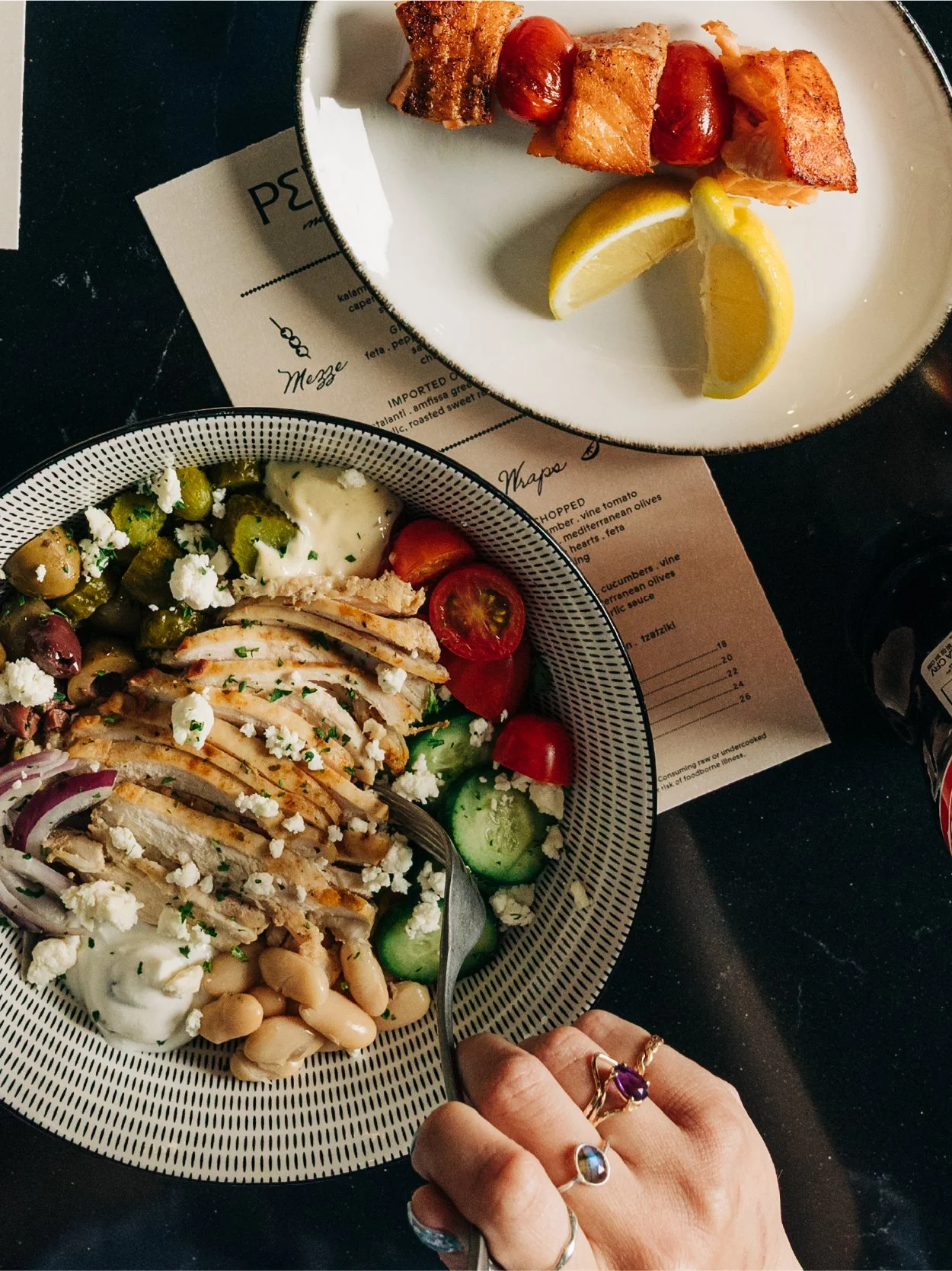 Plate with grilled chicken, cherry tomatoes, cucumber, and feta cheese salad. Another plate contains bacon-wrapped hot dogs with lemon wedges. A person wearing multiple rings is holding a fork over the salad.