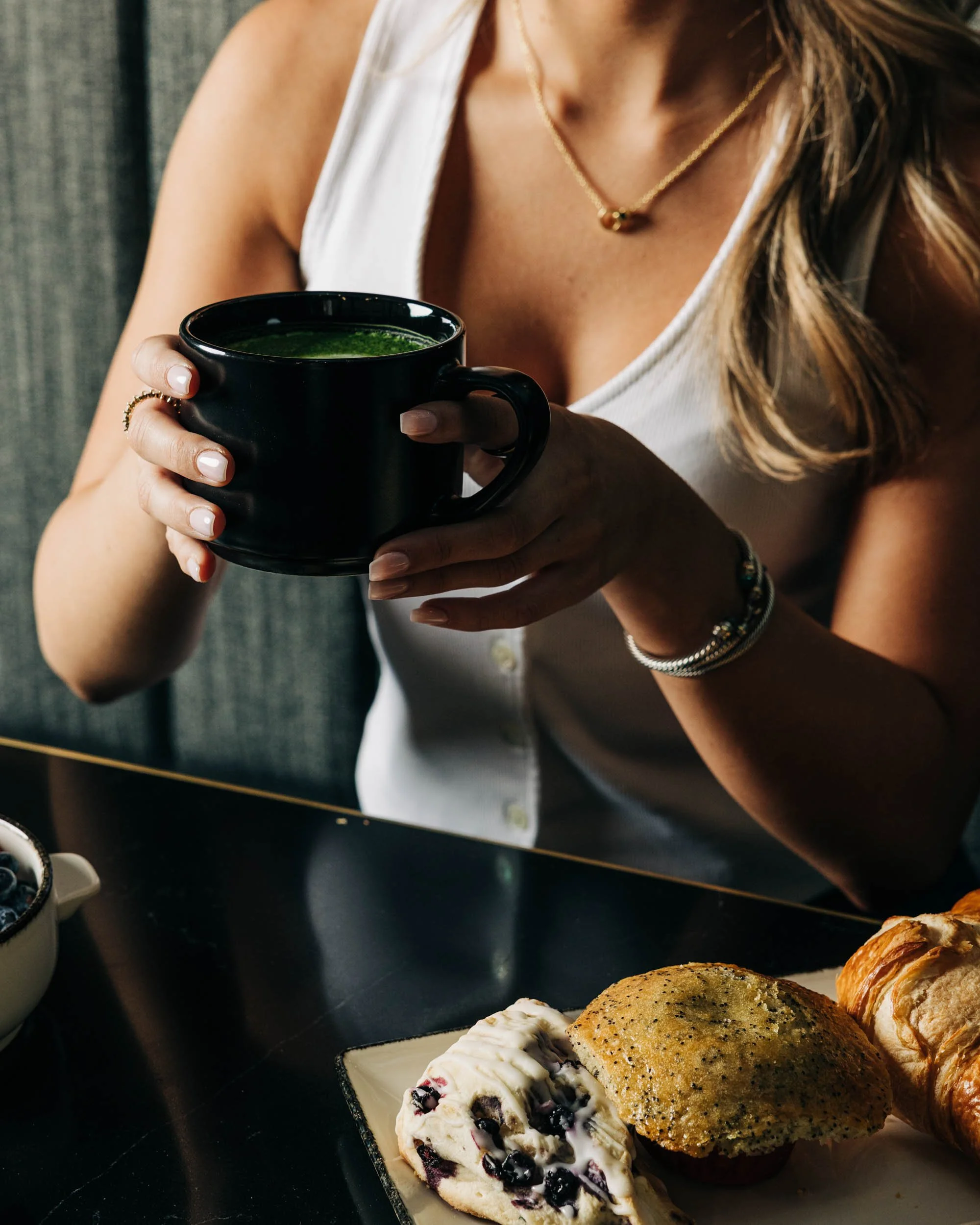 A woman holding a black coffee mug with a green beverage inside, sitting at a table with a plate of assorted pastries including a blueberry cheesecake, a cookie, and croissants.