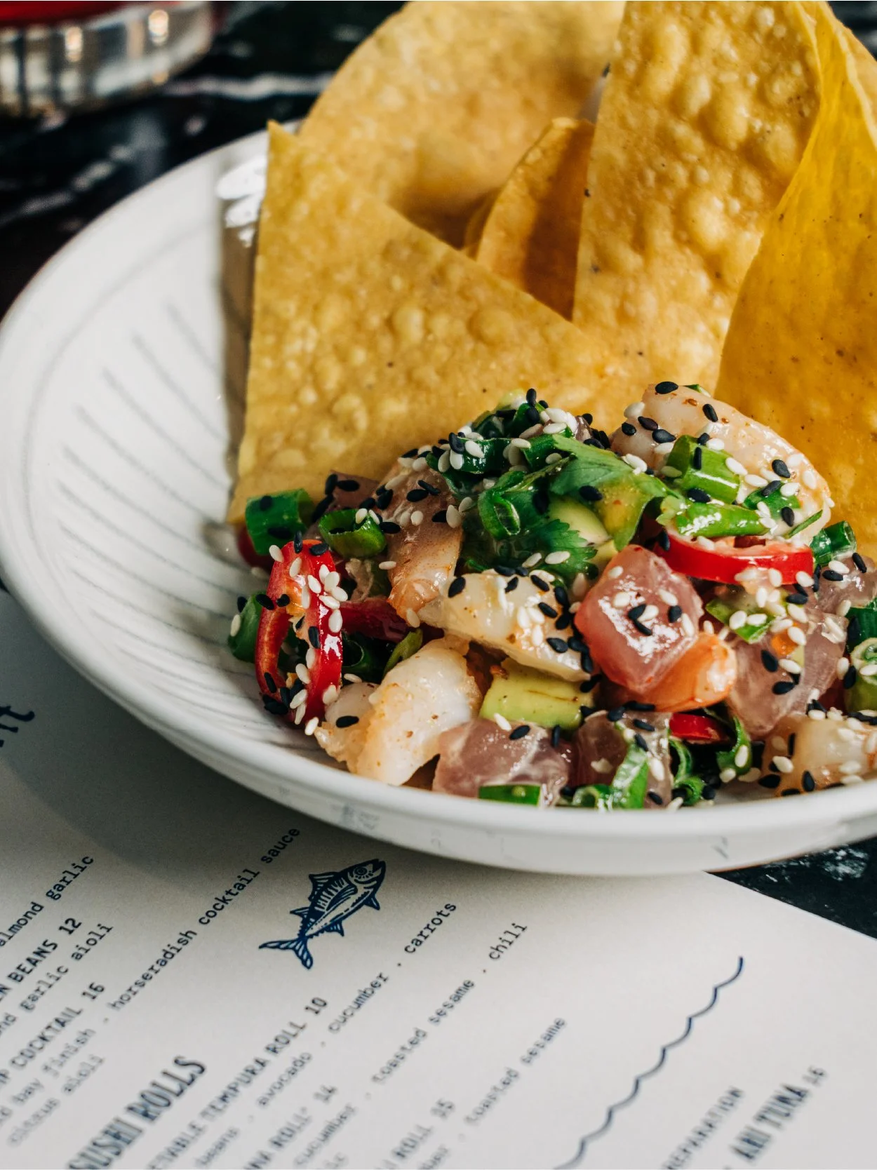 Bowl of shrimp and vegetable salad topped with sesame seeds and garnishes, accompanied by crispy tortilla chips, on a white plate with a menu underneath.