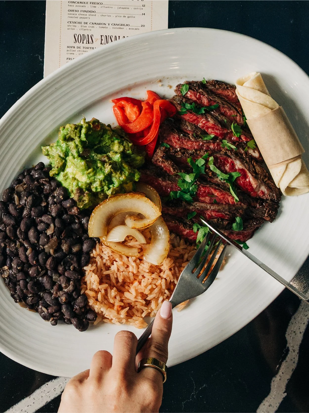 A white plate with grilled steak garnished with chopped cilantro, a serving of black beans, rice, grilled onions, guacamole, red peppers, and folded tortillas, with a fork and knife in the foreground, on a dark table with a menu in the background.