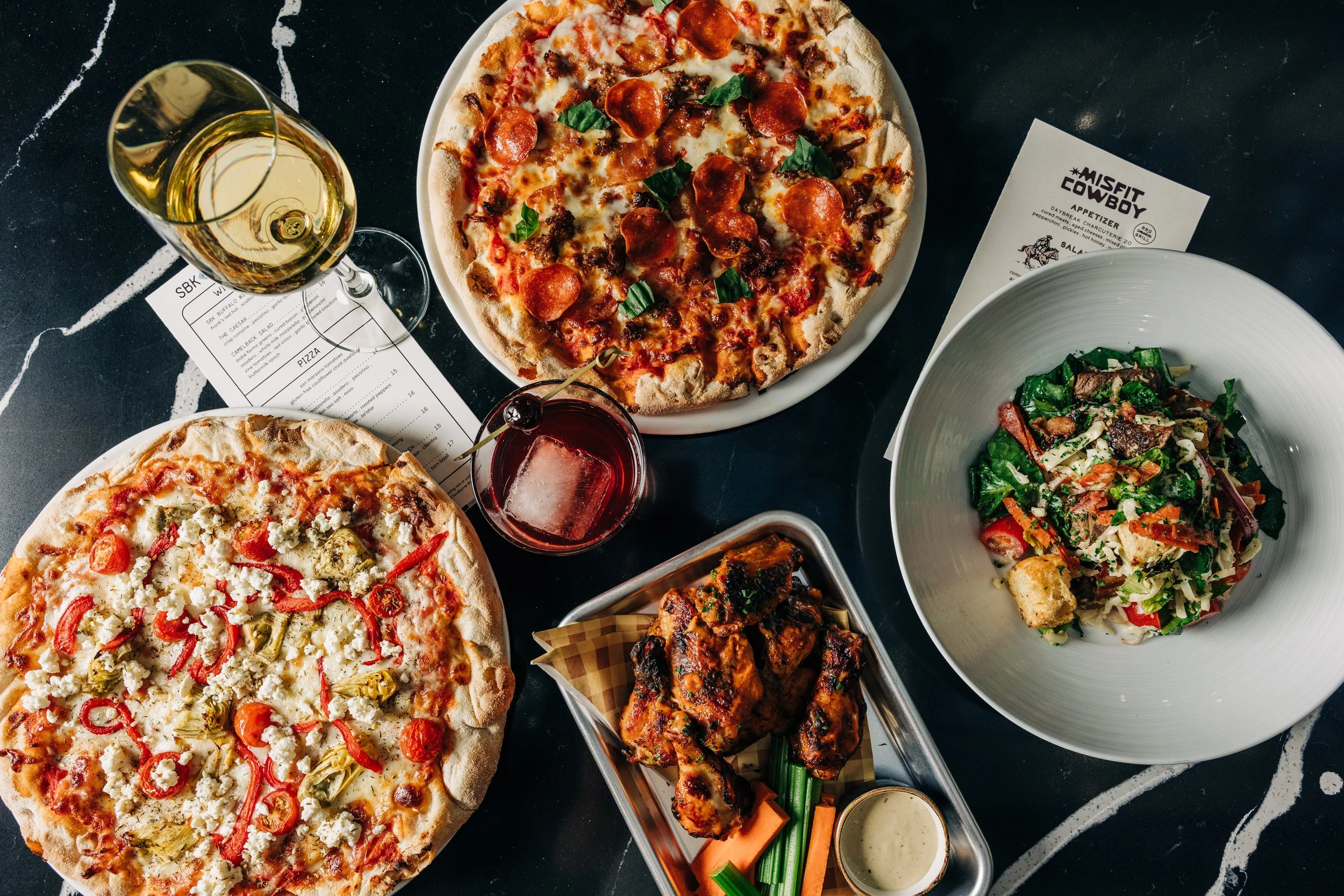 Assorted food on a black table including two pizzas with different toppings, a salad in a white bowl, a tray of grilled chicken wings, a glass of white wine, a glass of red drink with a cherry, and printed menus.