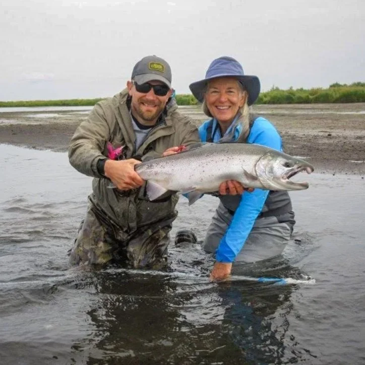 Two people, a man and a woman, standing in shallow water holding a large fish, smiling at the camera on a cloudy day with a grassy shoreline in the background.