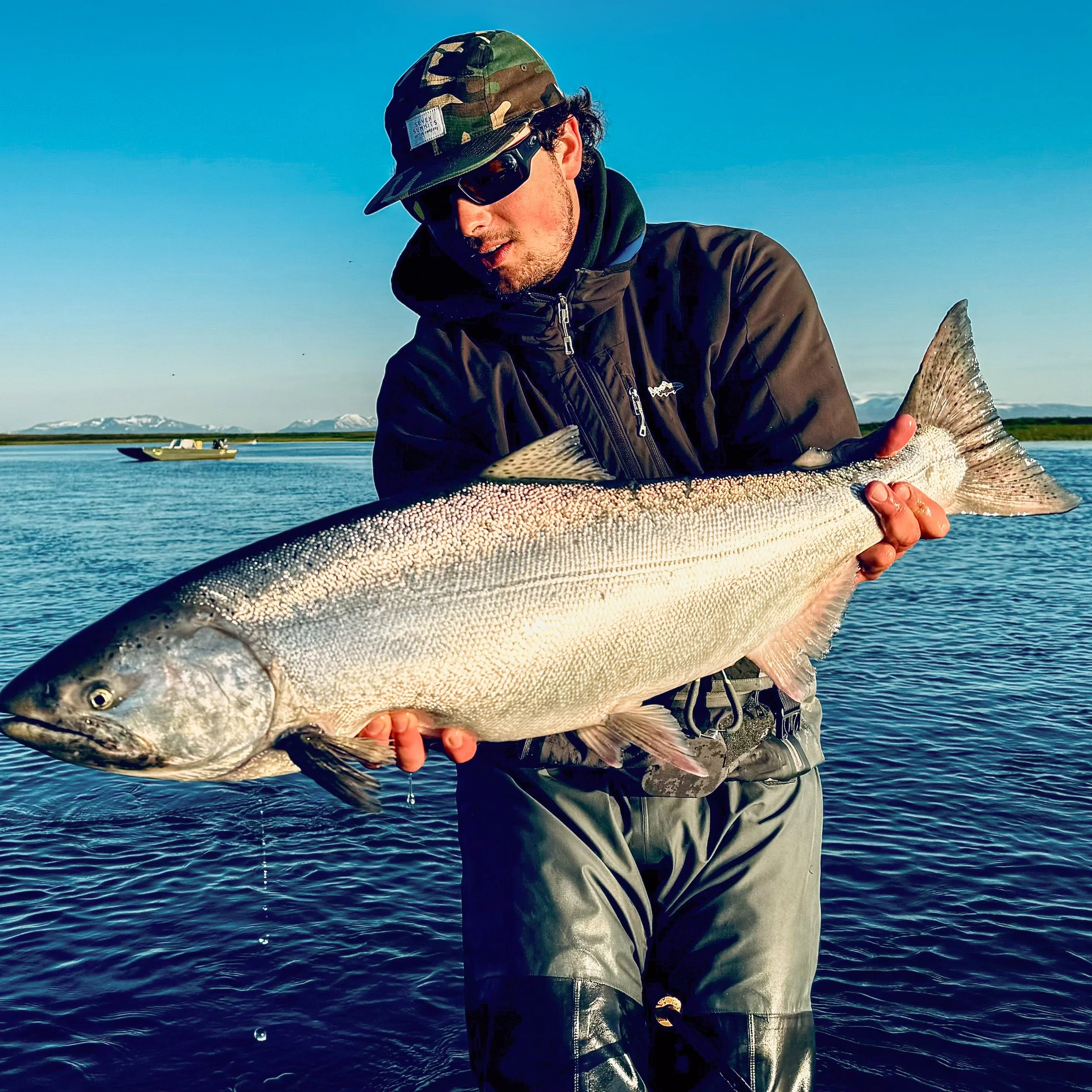 Person wearing a camouflage hat, sunglasses, and dark outdoor clothing, holding a large fish by the water with a boat and mountains in the background.