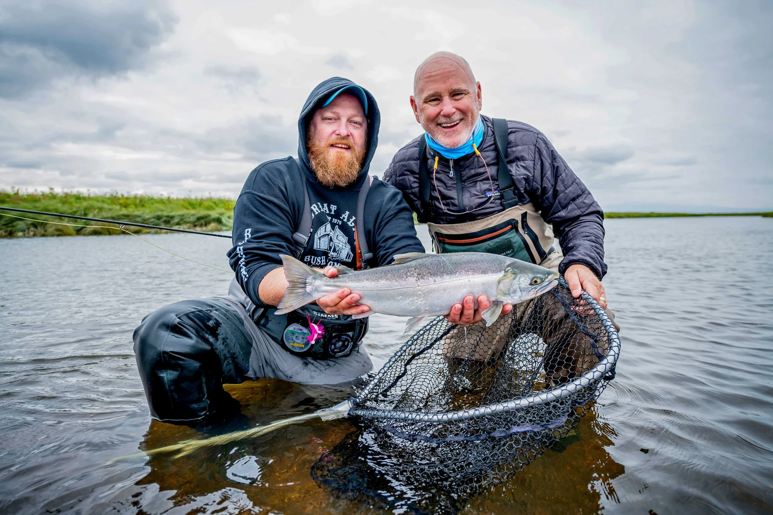 Two men, one with a beard and hoodie, the other bald with a beard, smiling and holding a large fish over water, with a net in the foreground and a cloudy sky in the background.