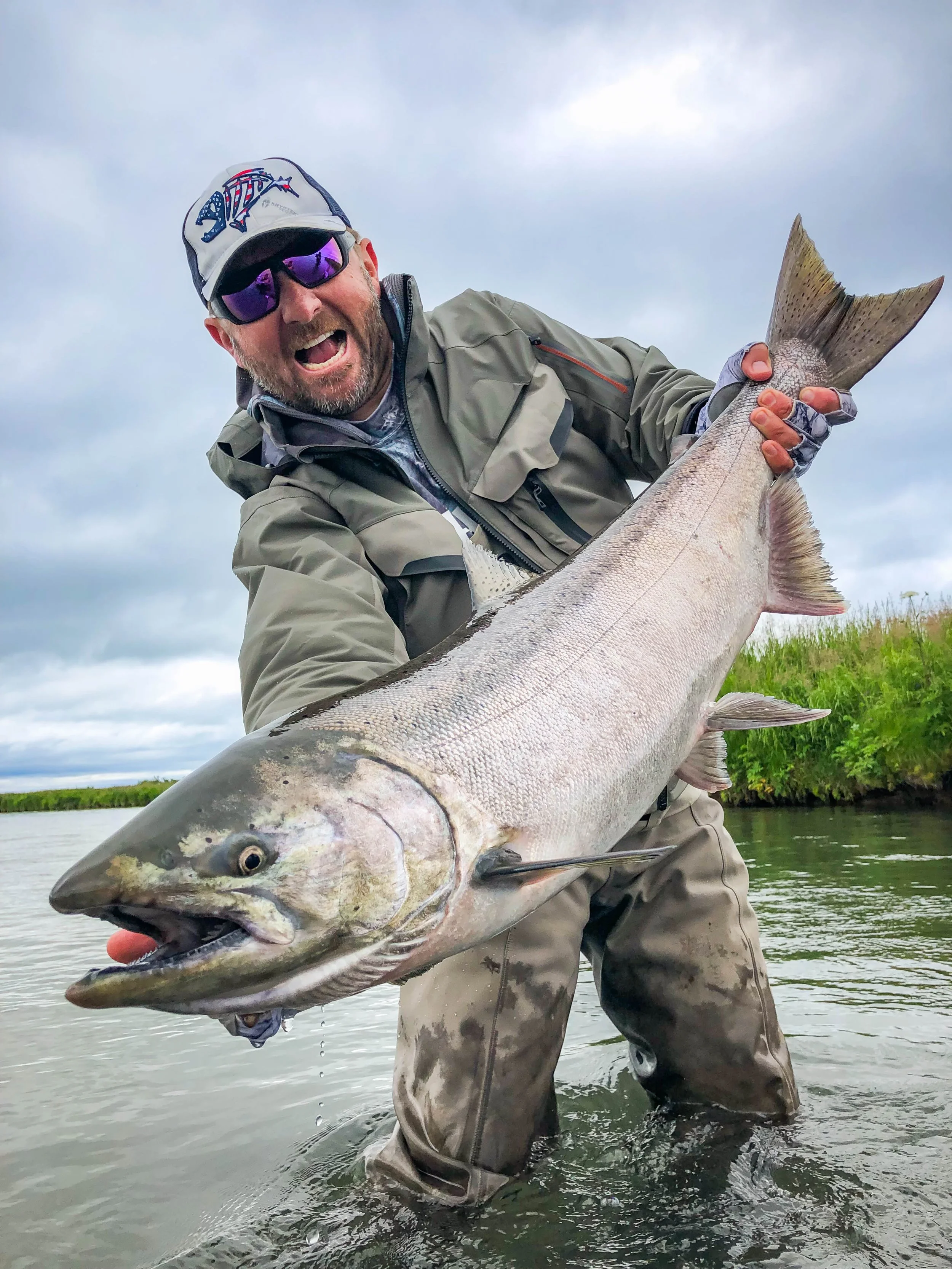 Man wearing sunglasses, a hat, and outdoor gear holding a large fish in shallow water with a cloudy sky and green vegetation in the background.